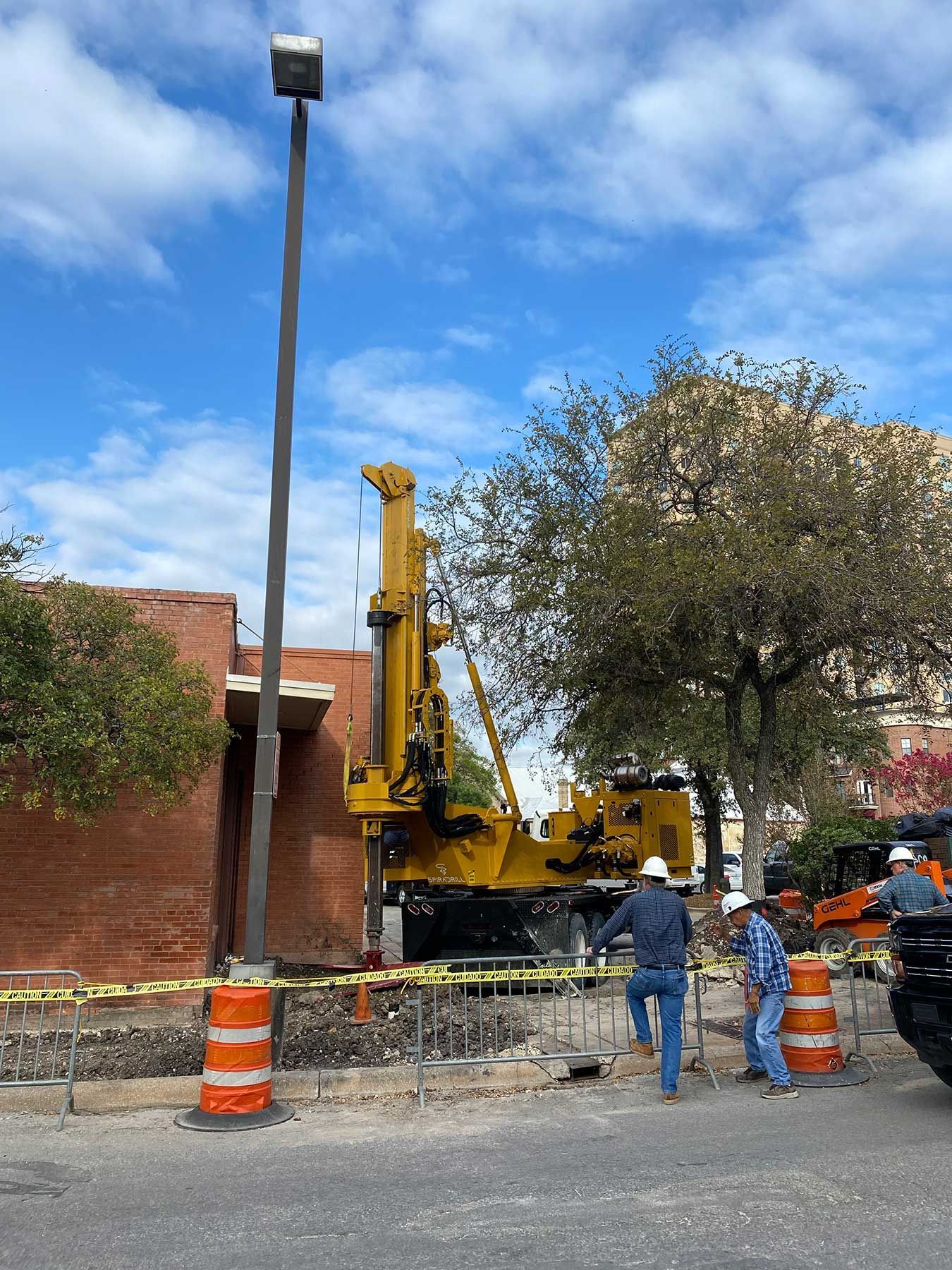 A yellow drilling rig operates at a construction site near a brick building and a tall lamp post, with workers nearby.