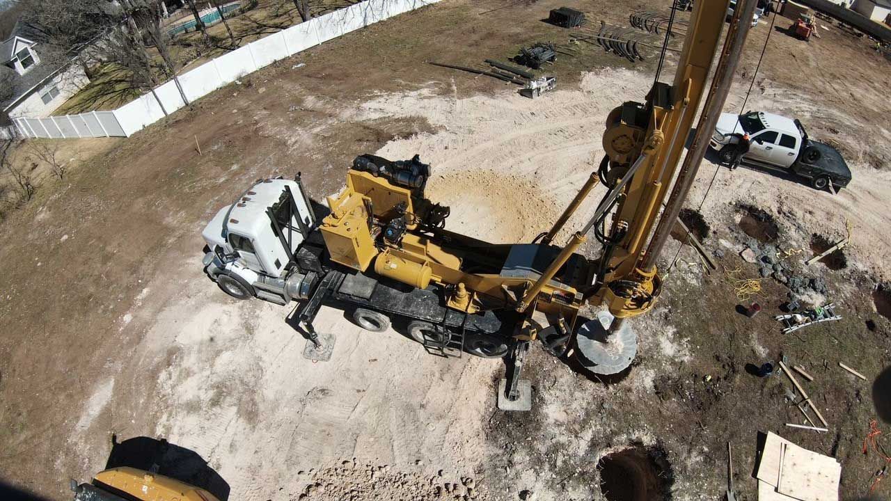 Aerial view of a yellow truck-mounted drilling rig operating on a construction site next to a white pickup truck.