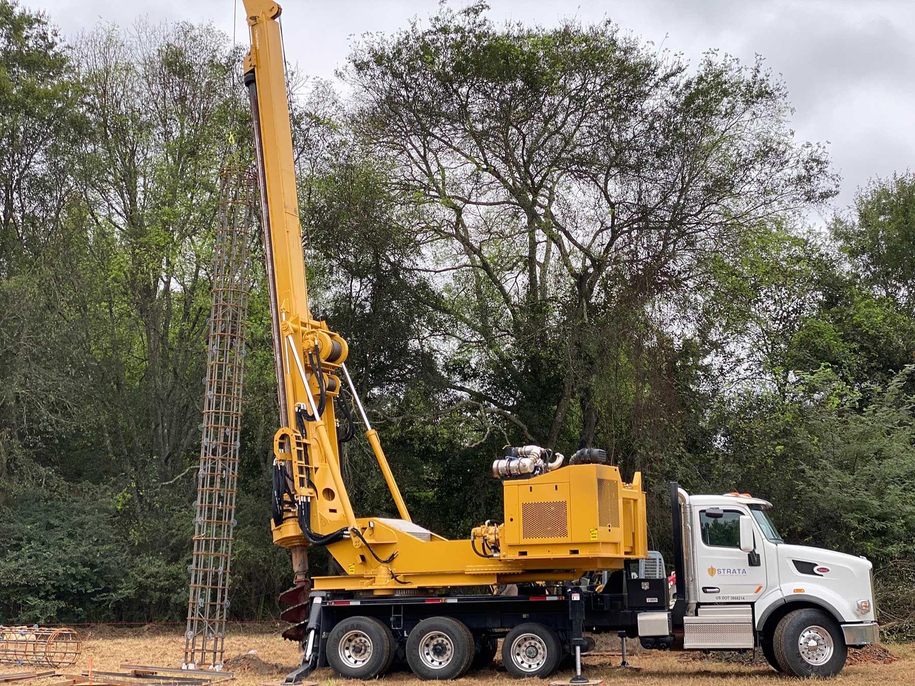 A yellow drilling rig mounted on a white truck parked in front of trees, holding a metal rebar cage vertically.