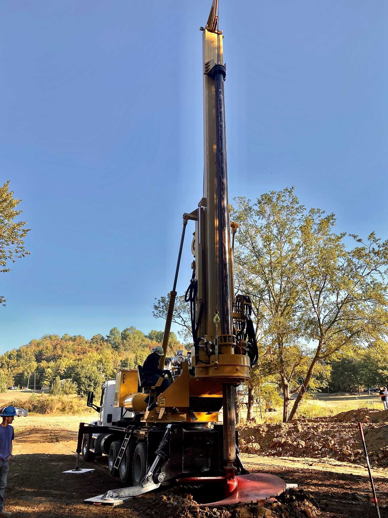 A yellow truck-mounted drilling rig operating in a rural, sunny setting with trees in the background.