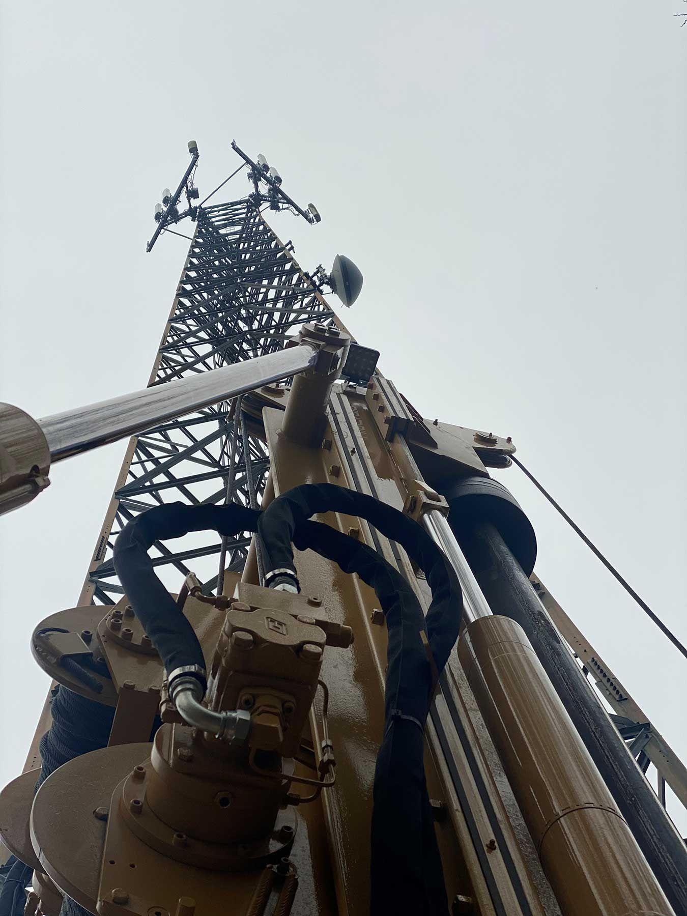 A low-angle view of a large, brown industrial drill rig positioned in front of a tall, metal communications tower.