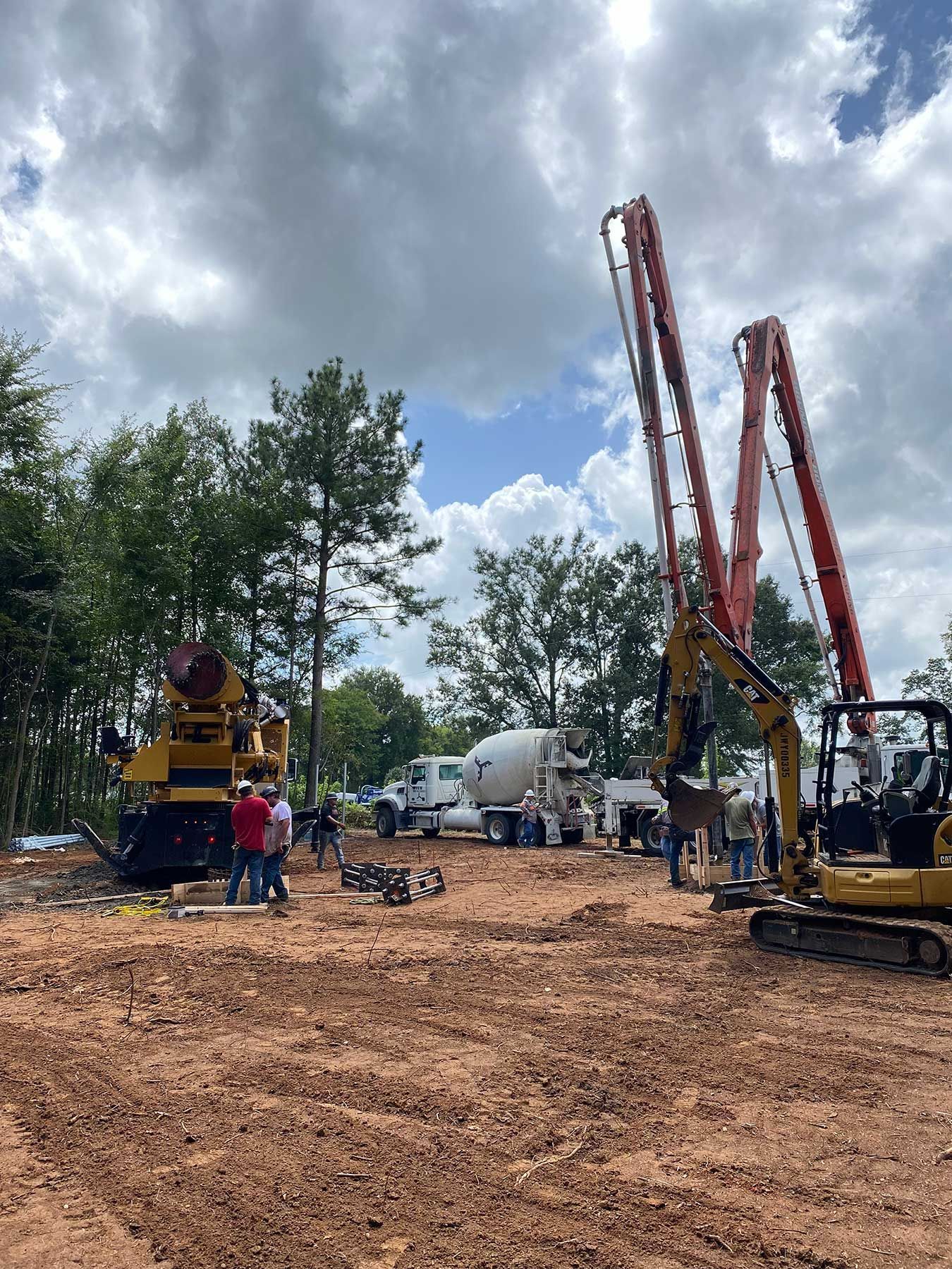 Construction crew and machinery, including a concrete mixer and a boom pump, on a dirt site under a cloudy sky.