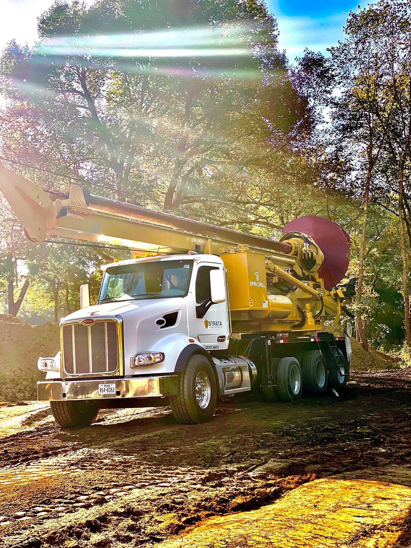 A white industrial drill truck parked on a dirt site in front of a sunlit forest.