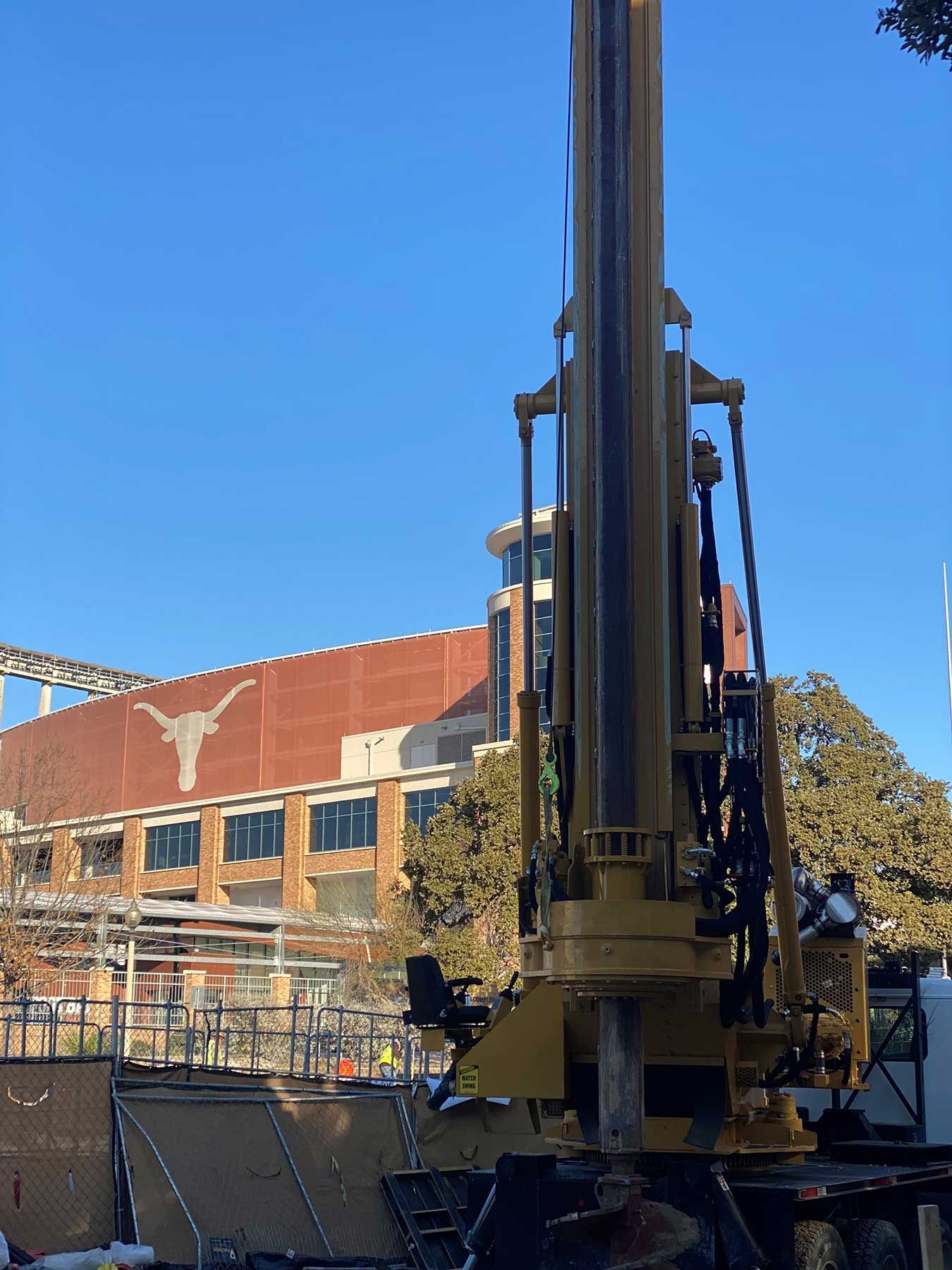 A yellow drill rig stands in front of a brick building featuring a large, white Texas Longhorn logo under a blue sky.