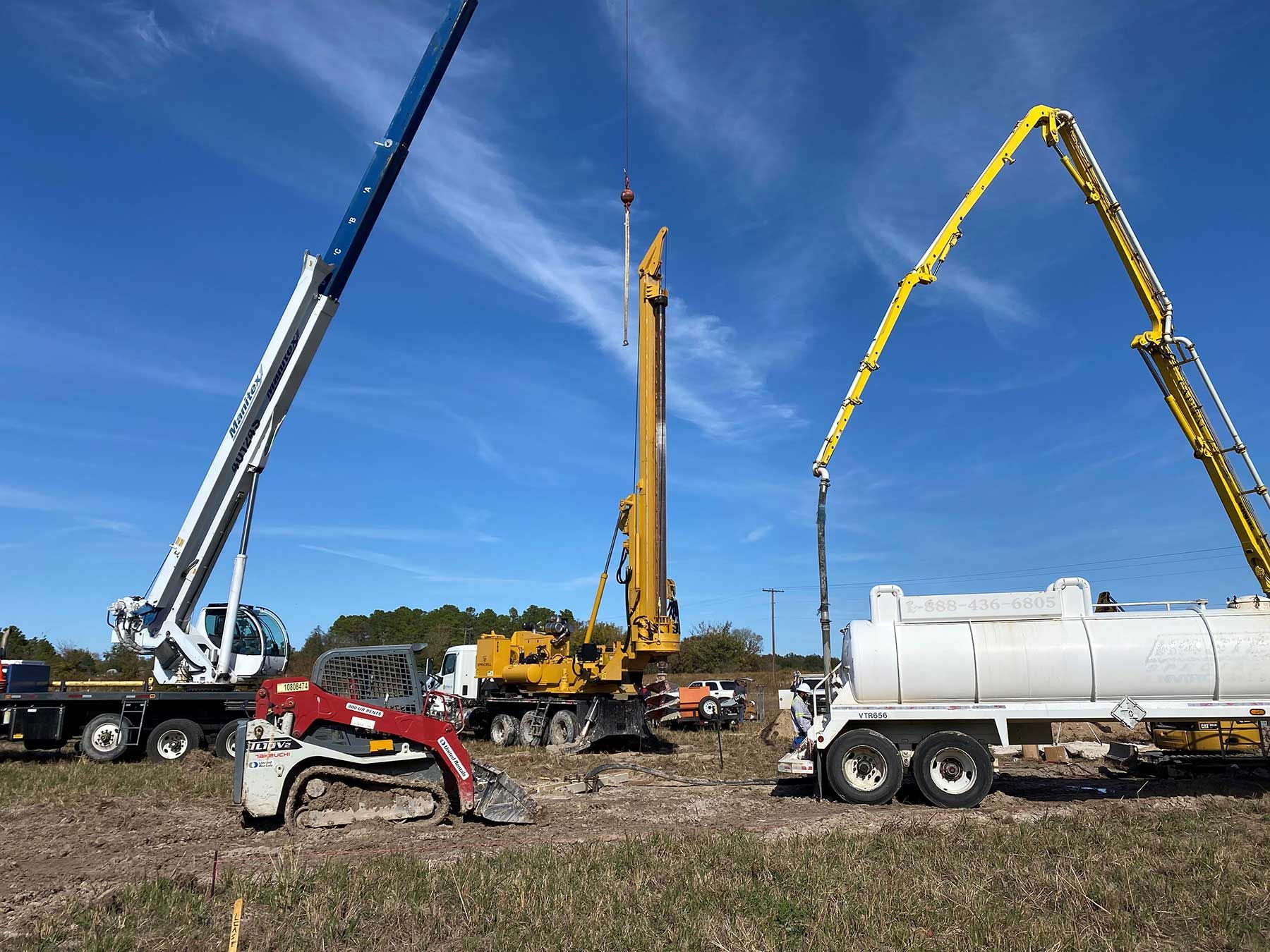 Construction site with a white crane, a yellow drill rig, a red skid steer, and a yellow concrete pump truck.