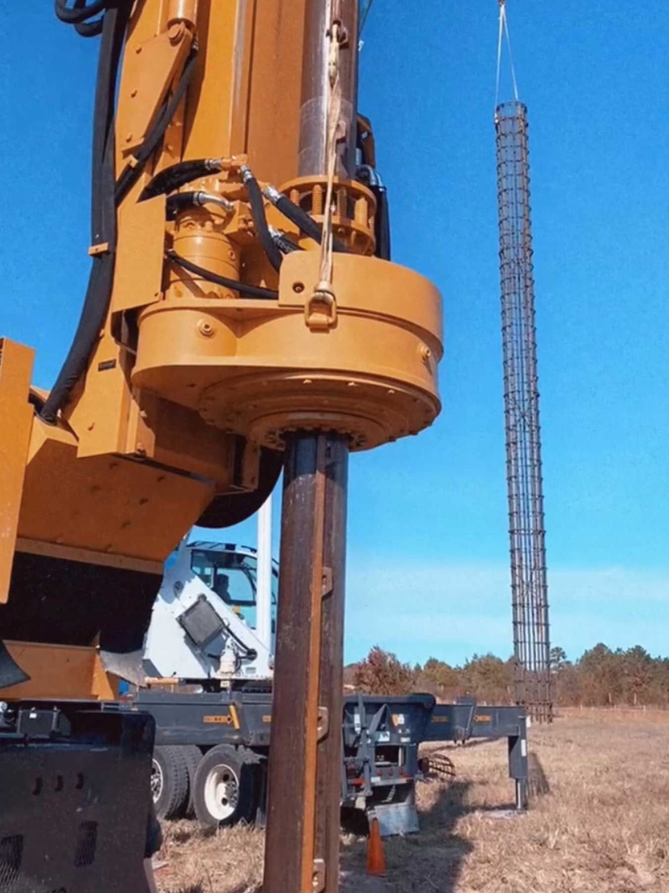 A yellow rotary drilling rig on a construction site, with a suspended metal rebar cage in the background.