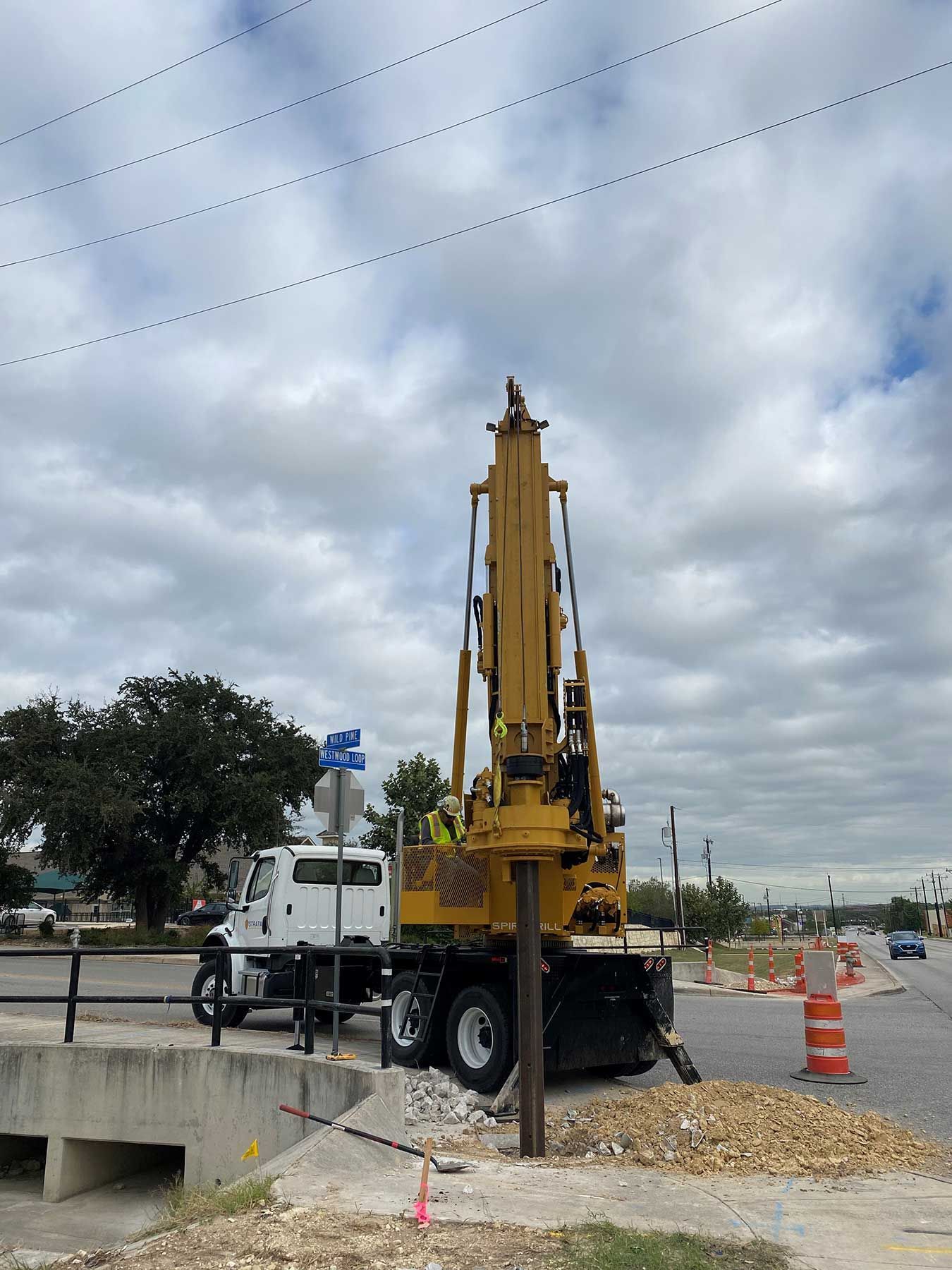 A yellow construction vehicle with an upright drilling attachment positioned near a street corner and a concrete culvert.