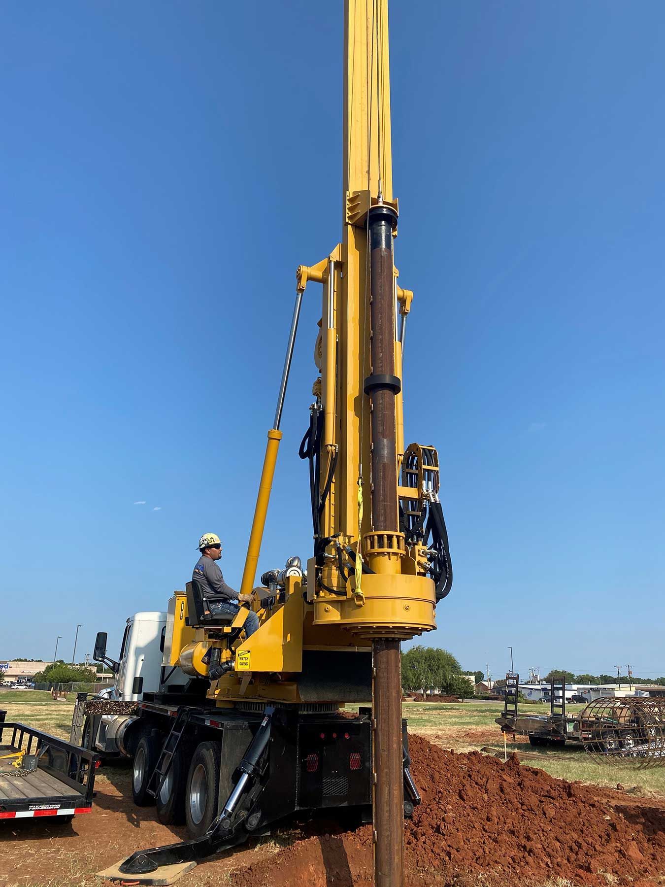 A yellow drill rig sits on a construction site, driving a metal pile into the ground under a clear blue sky.