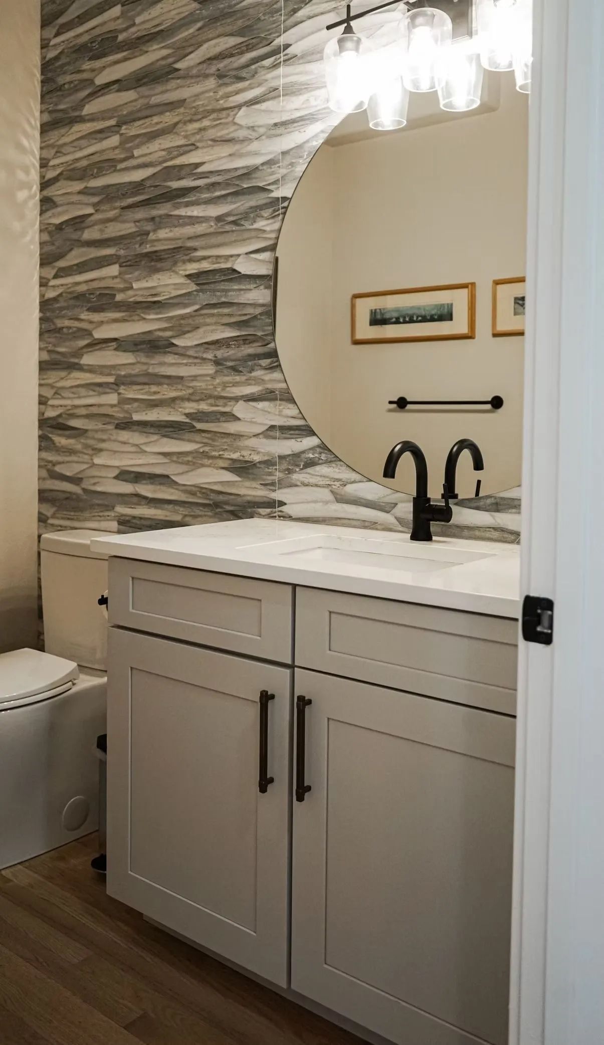 A small bathroom with a gray stone accent wall and a round mirror above a white cabinet. Black fixtures and framed art are visible.