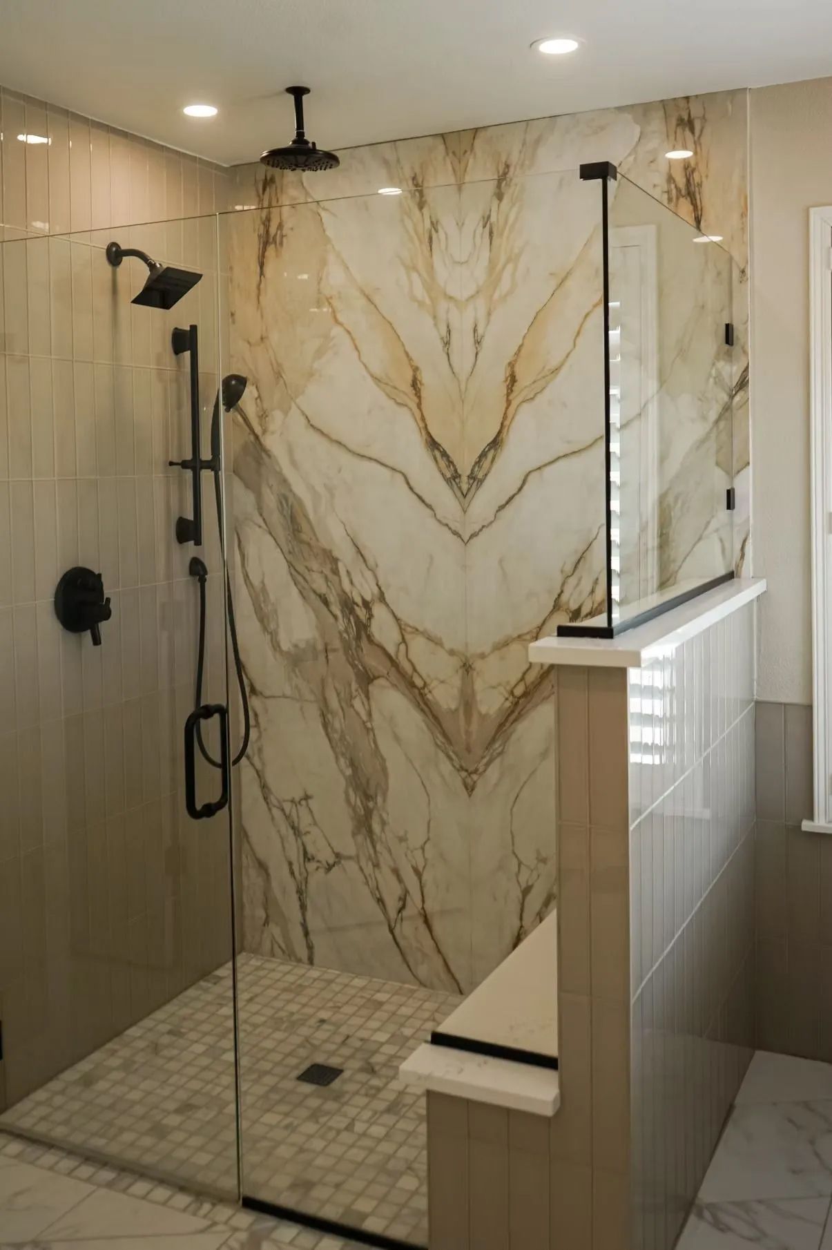 A modern bathroom shower with large marble-like tiles and glass enclosure. Features black shower fixtures and a low wall with a countertop.