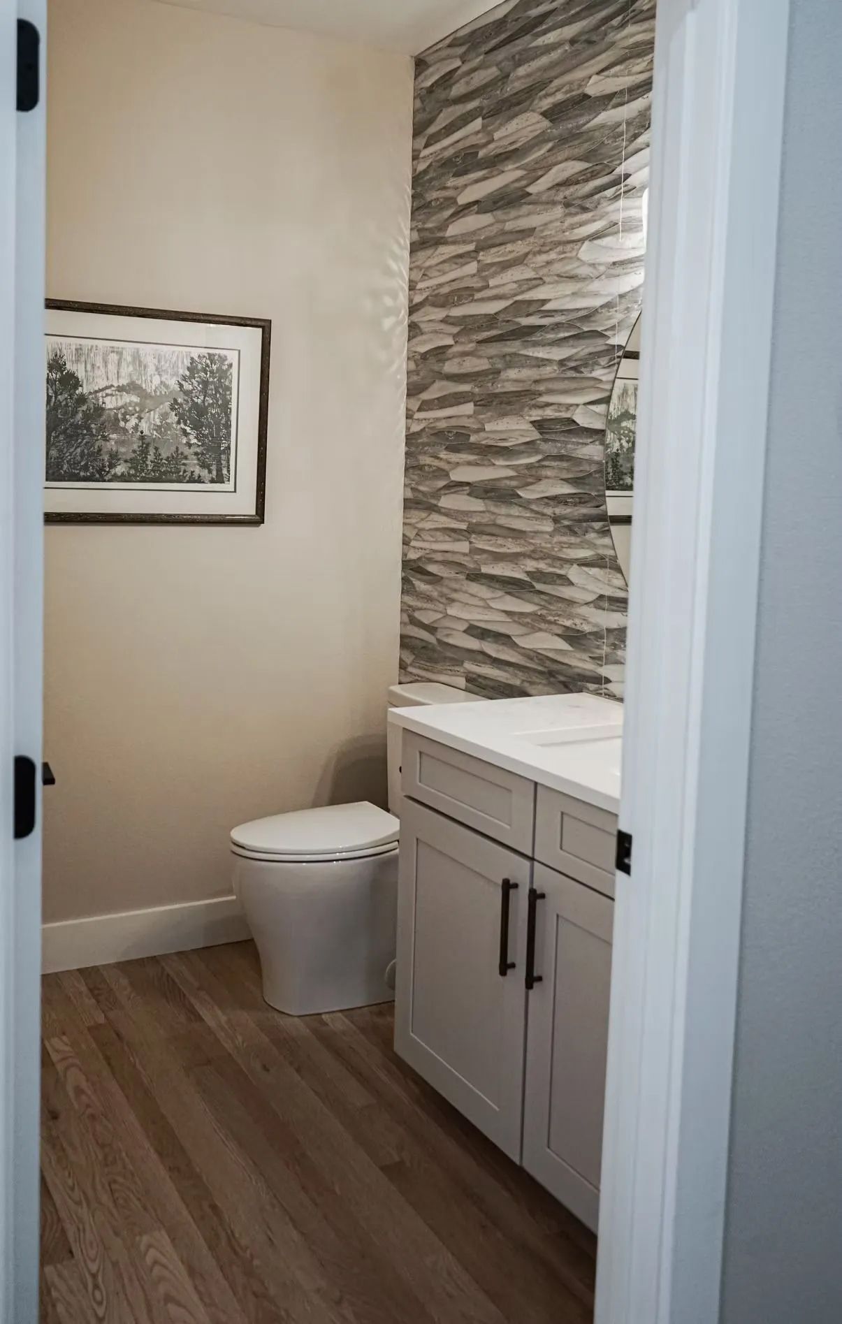 A small, well-lit bathroom with a white toilet, vanity, and a textured stone accent wall.  A framed picture hangs on a beige wall.