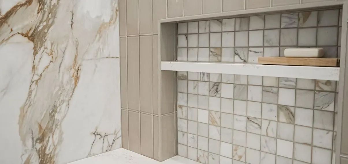 Shower with marble-like patterned wall tiles and a built-in niche with small square tiles and a shelf holding soap and a wooden tray.
