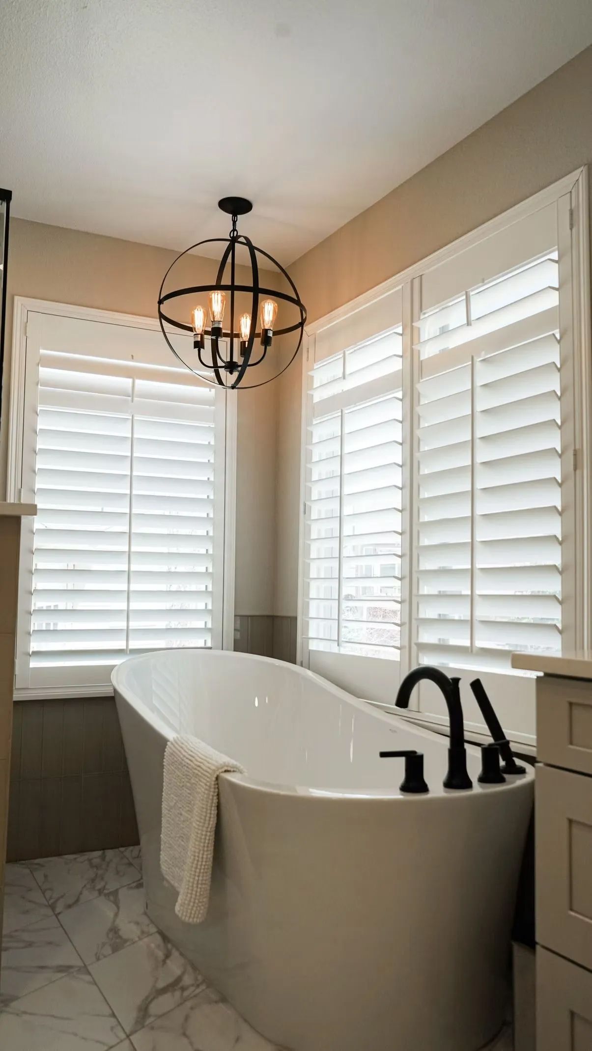 Freestanding white bathtub in a bathroom with white shutters, black fixtures, and a dark chandelier.