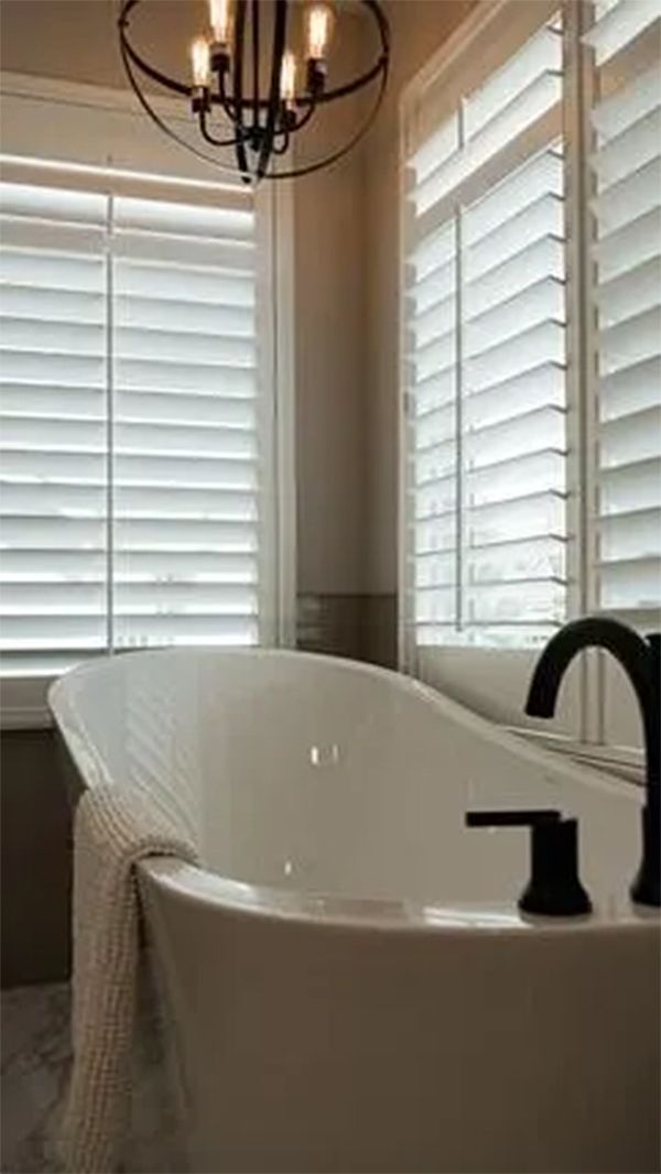 Freestanding white bathtub in a bathroom with white shutters, black faucet, and a decorative light fixture.