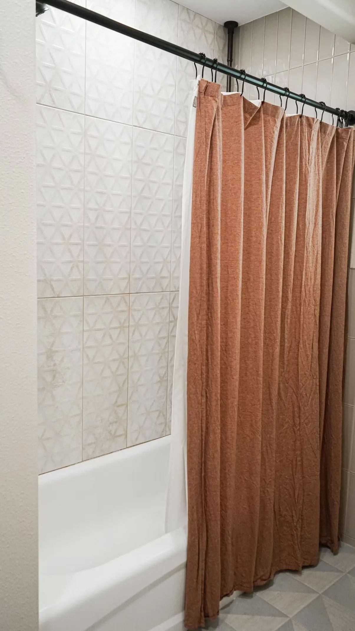 A bathroom with a white bathtub, patterned tile, and a rust-colored shower curtain. A black shower rod is installed.