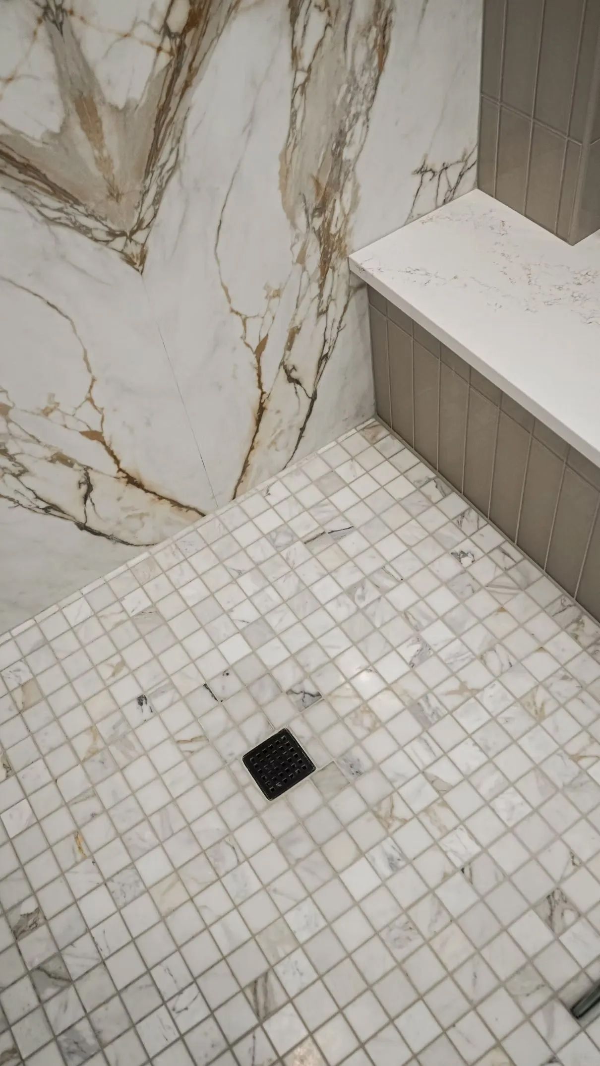 Shower with marble tile floor and walls, featuring a black drain and a white bench.
