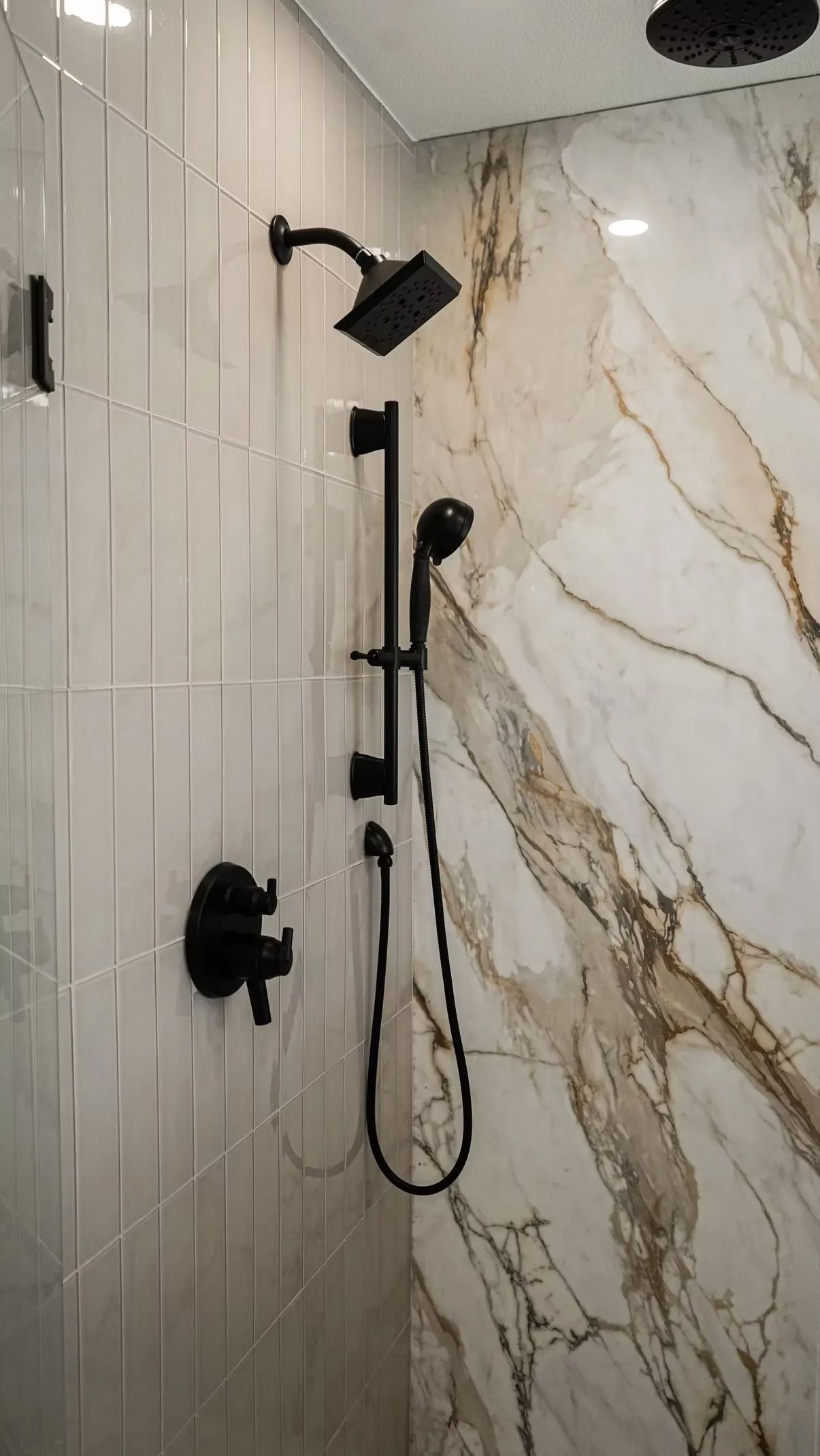 Black shower fixtures on a marble-look tiled shower wall. A handheld showerhead and a rain showerhead are visible.