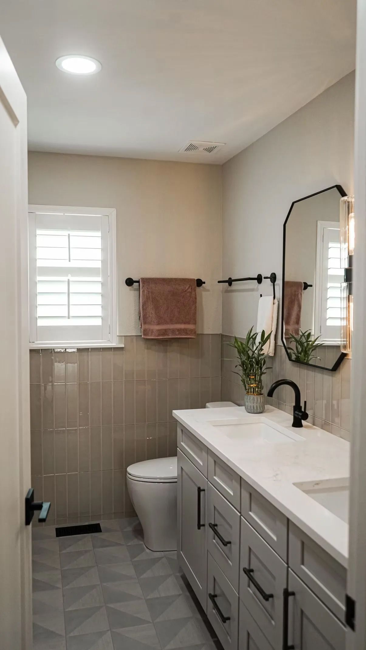 Bathroom with white vanity, grey tiled floor, and tan walls. Features include a toilet, window with shutters, and black fixtures.