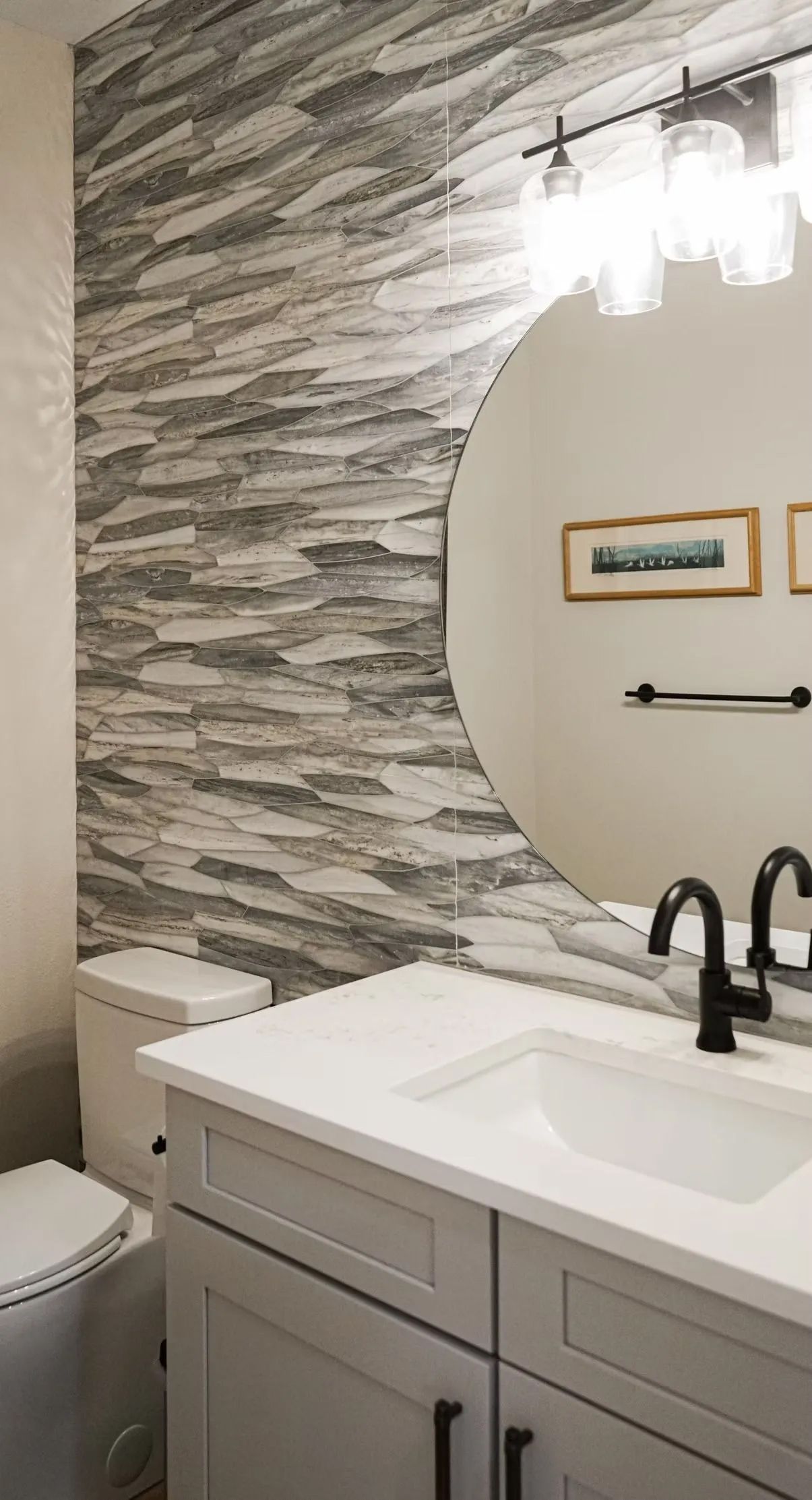 Bathroom with gray stone accent wall, white vanity, round mirror, and black fixtures.