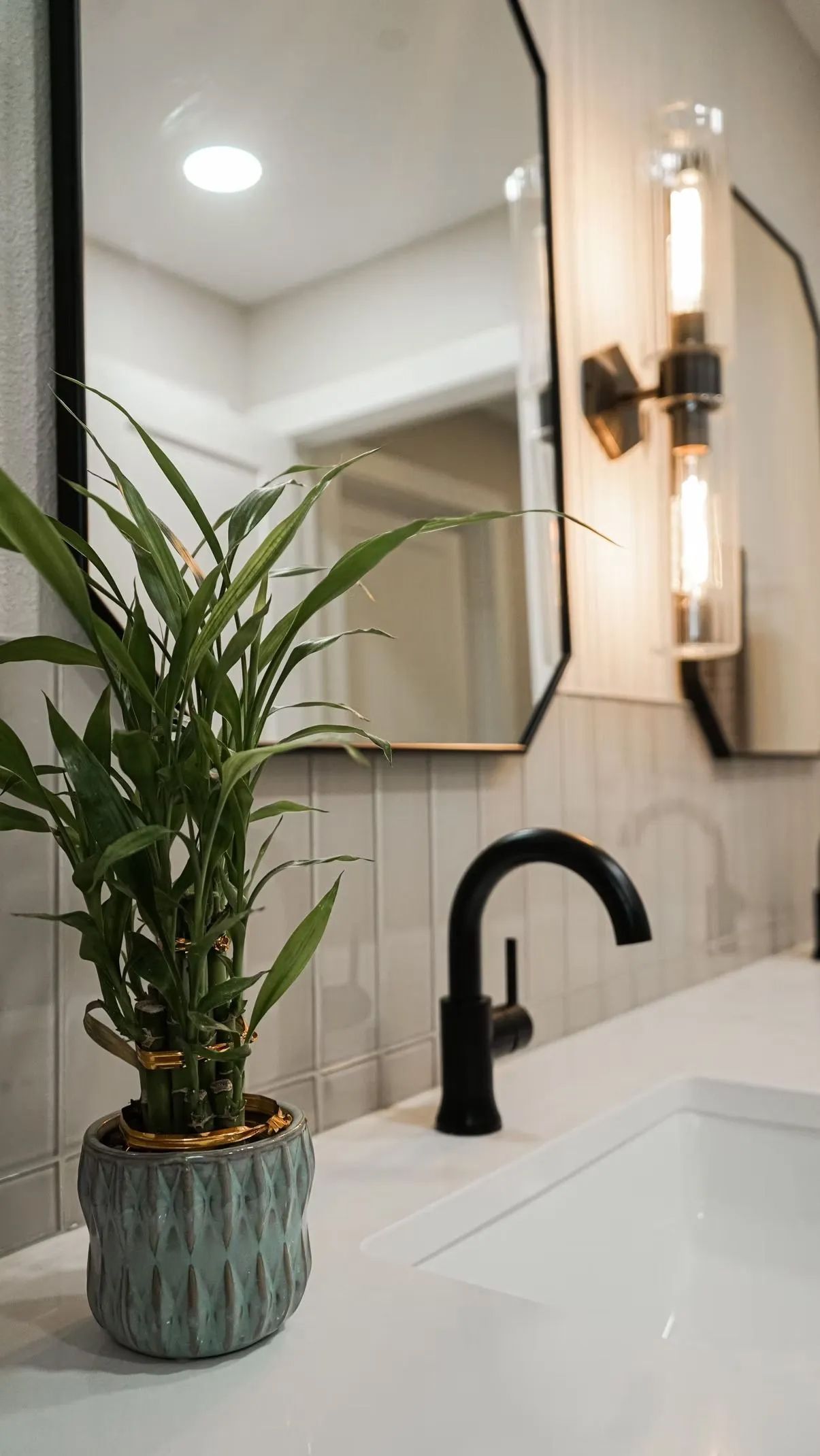 Bathroom with a black faucet, white countertop, and a potted bamboo plant. A decorative mirror and sconce are visible.