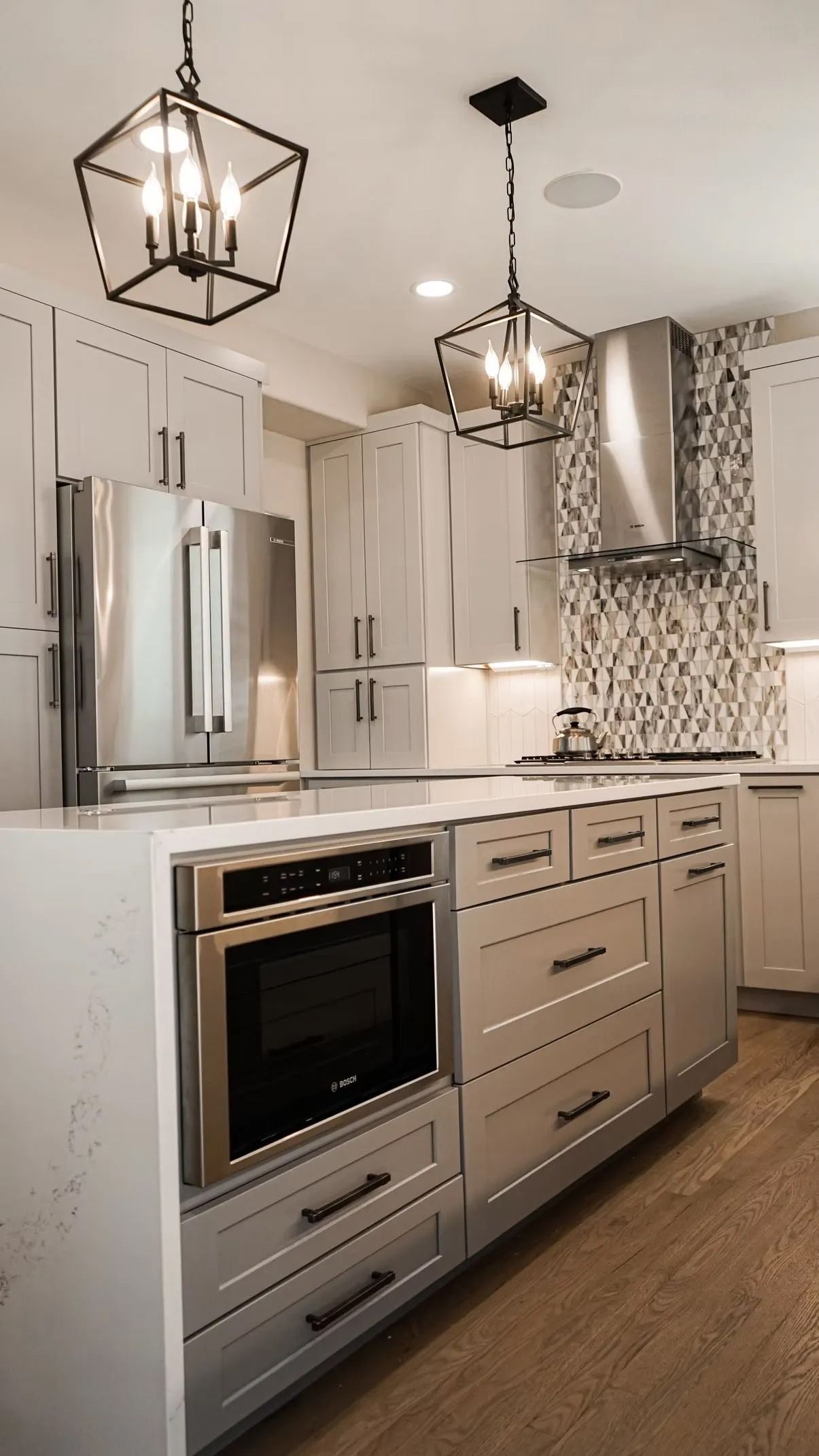 Modern kitchen with a white island, built-in oven, and stainless steel appliances. Geometric light fixtures hang above.