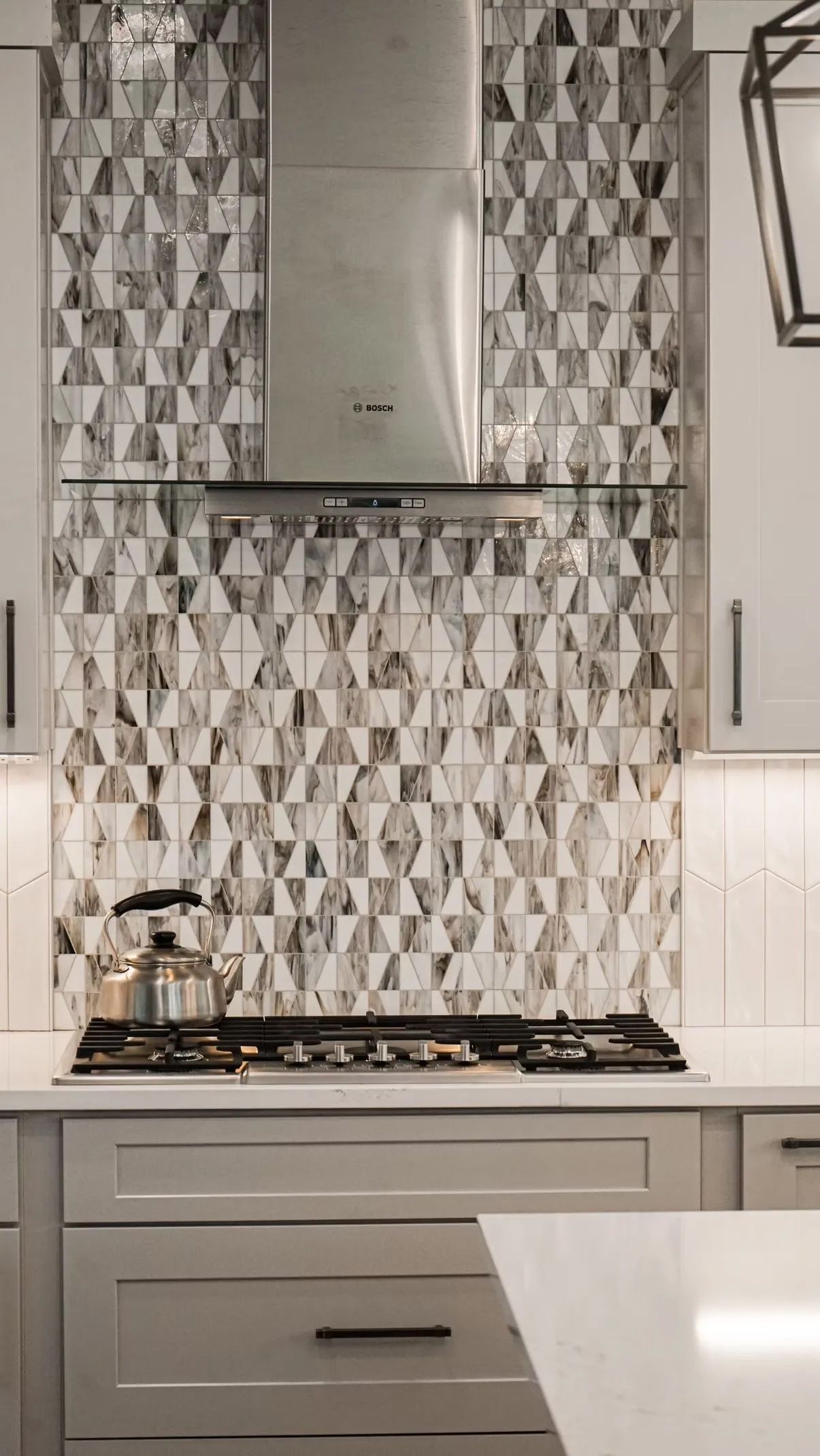 Kitchen with stainless steel range hood and geometric tile backsplash above gas stovetop, light gray cabinets.