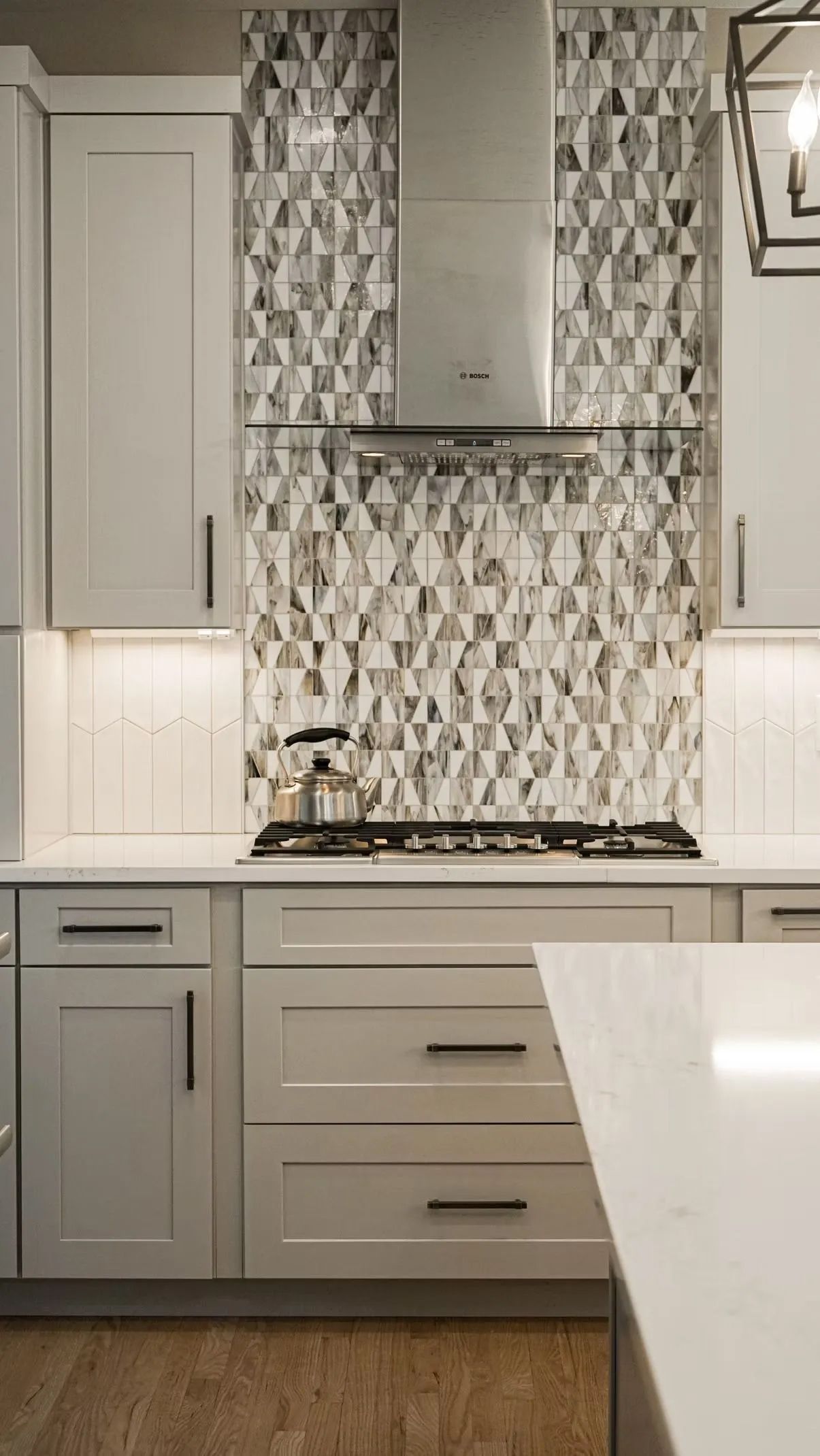 A modern kitchen with light gray cabinets, white countertops, and a mosaic backsplash.  A stainless steel range hood is centered above the stove.