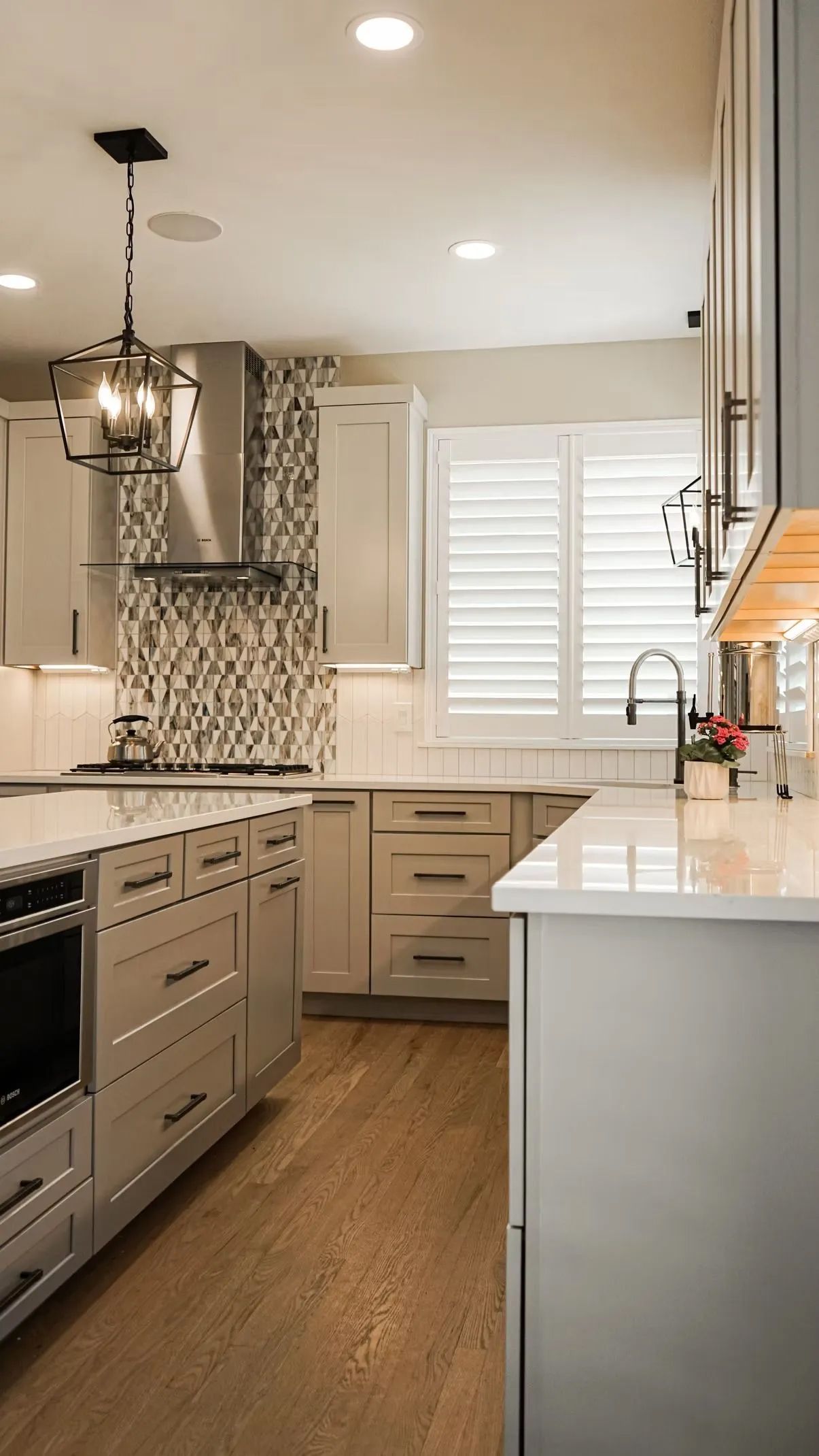 A modern kitchen with light gray cabinets, stainless steel appliances, and hardwood floors. White countertops and a window with shutters provide natural light.