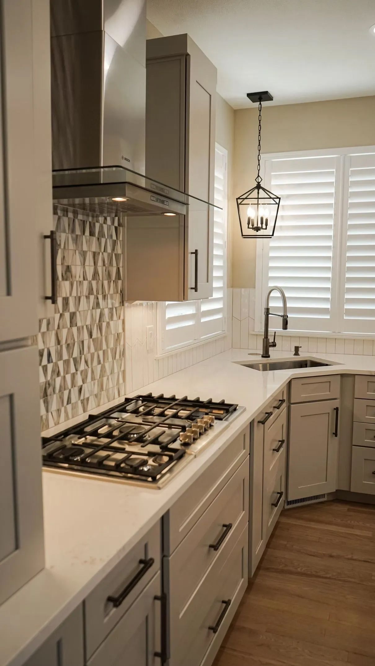A modern kitchen with white cabinets, a gas stovetop, and a stainless steel range hood. Light floods in from a window with white shutters.