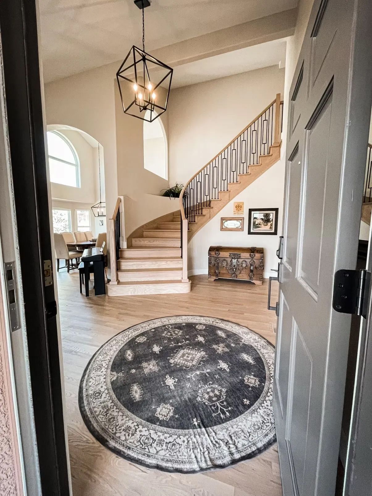 A bright entryway with a round rug, staircase, chandelier, and a glimpse of the dining area through a doorway.