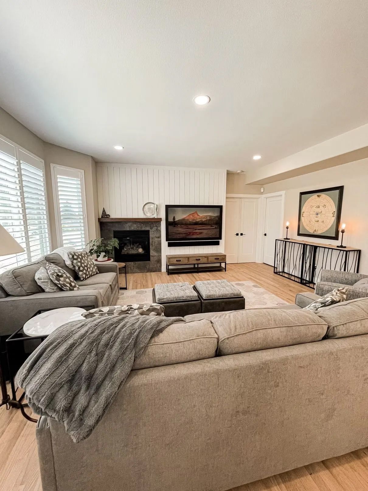 Spacious living room with neutral tones, featuring a gray sectional sofa, fireplace, and large TV. Hardwood floors and a console table are also visible.