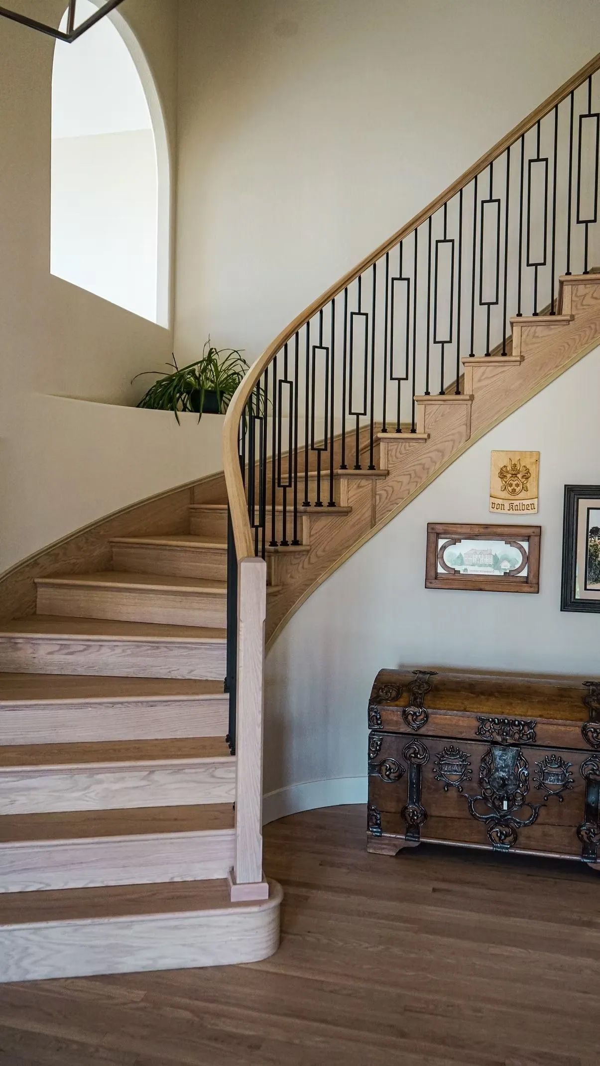 Wooden staircase with decorative metal railing ascends past an alcove with plants. A detailed wooden chest and framed artwork sit below.