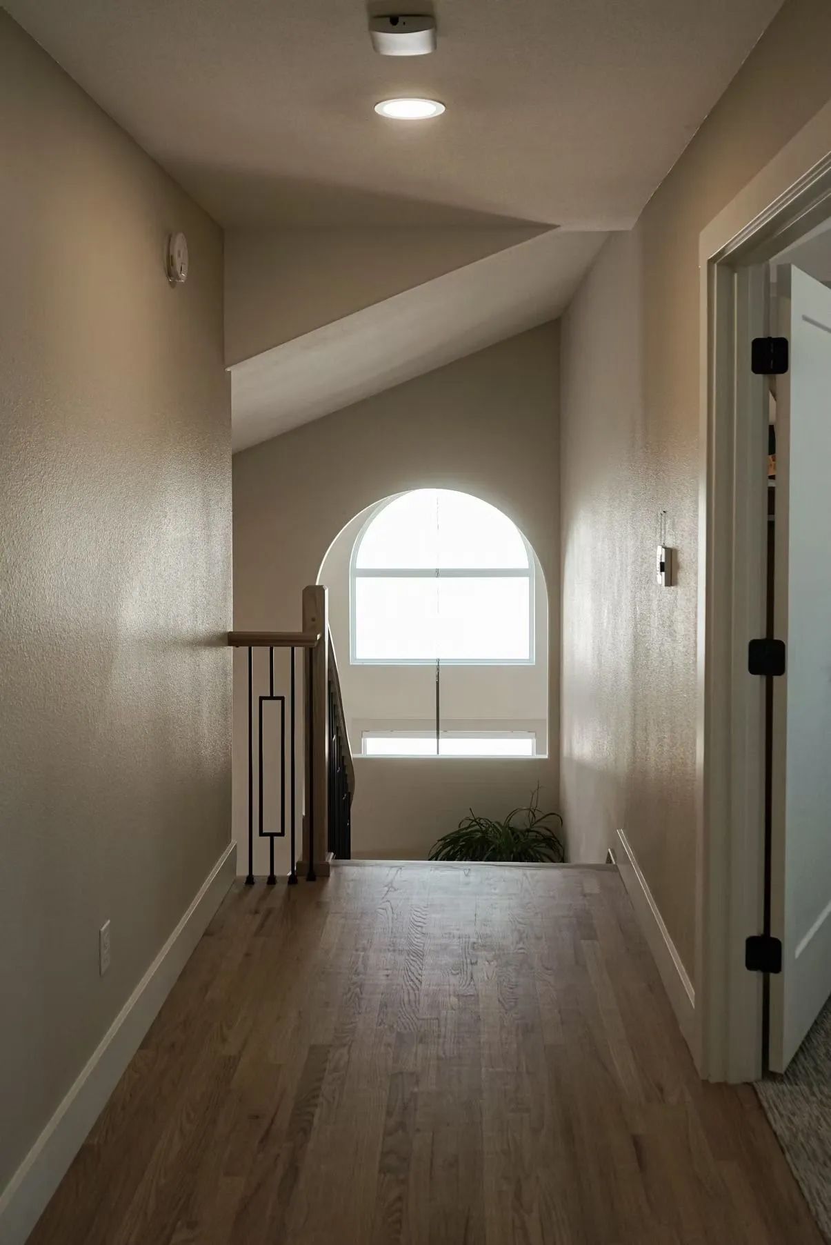 Hallway with hardwood floor leading to an arched window. Stairs and a black and white railing are visible to the left.