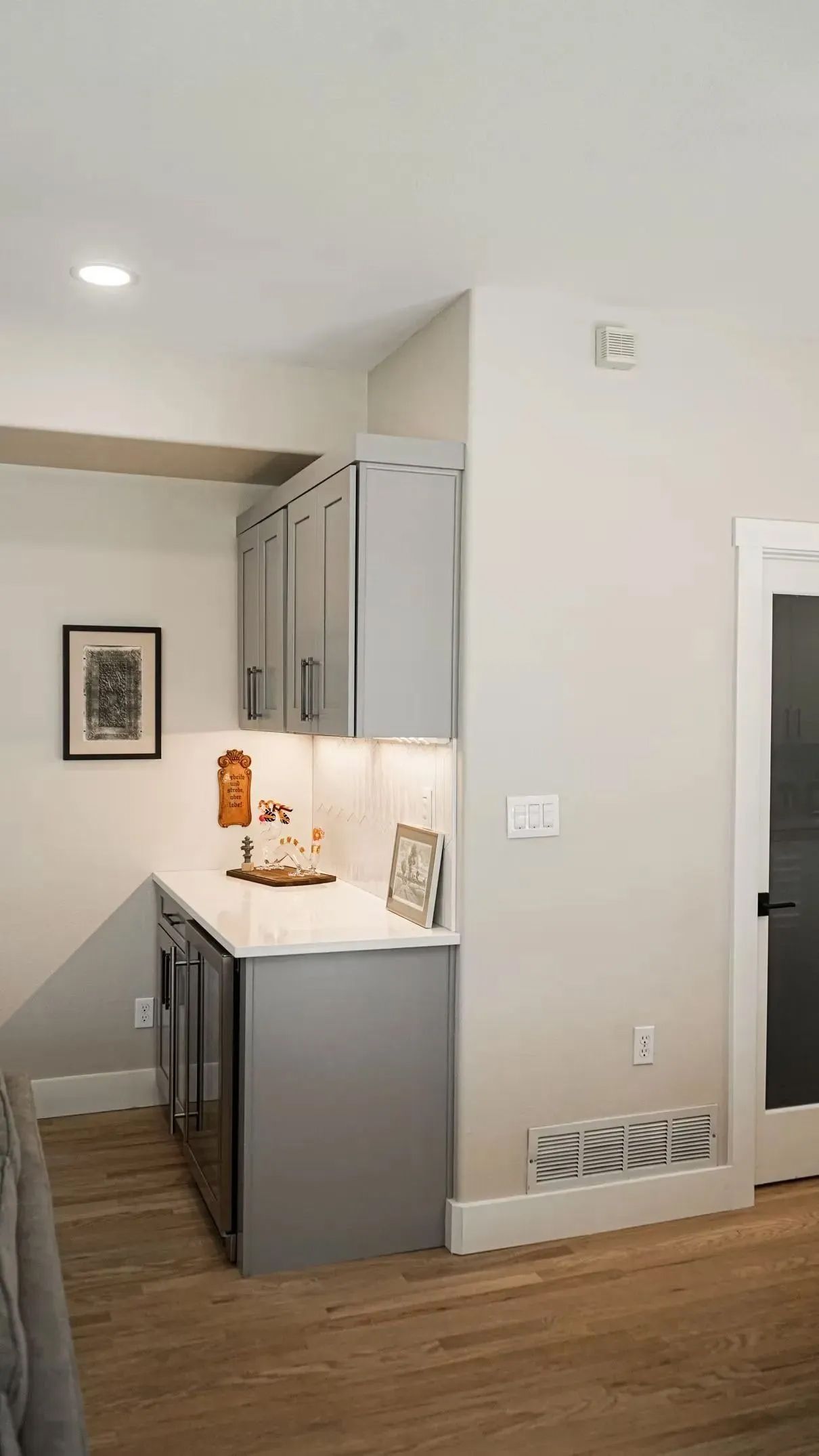 A small gray and white wet bar area with cabinets and a built-in mini-fridge; a framed picture hangs on the wall.