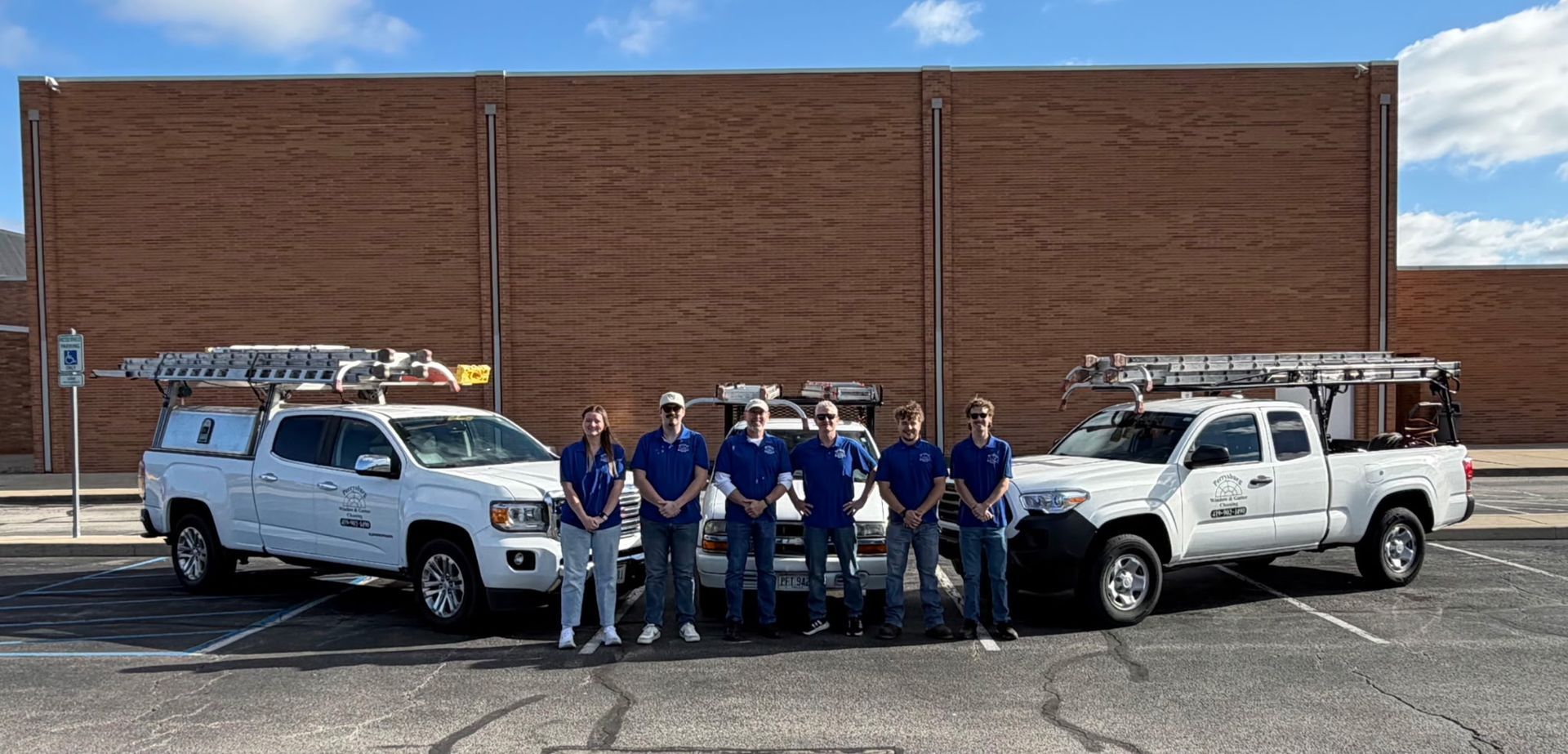 Six people in blue shirts stand in front of two white utility trucks with roof ladders parked against a brick wall.