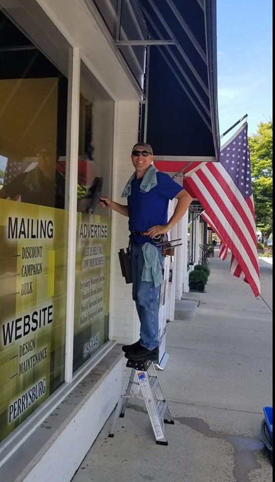 Man washing storefront window, standing on a step stool. American flag waving nearby.