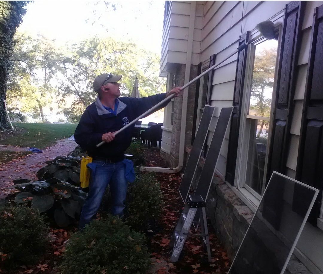 Man cleaning window of a house with a long-handled brush, fall foliage in the background.