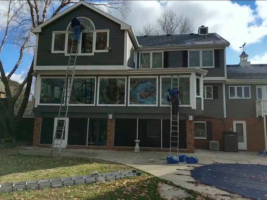 Two people on ladders painting a two-story house with dark gray siding. Sunny day.
