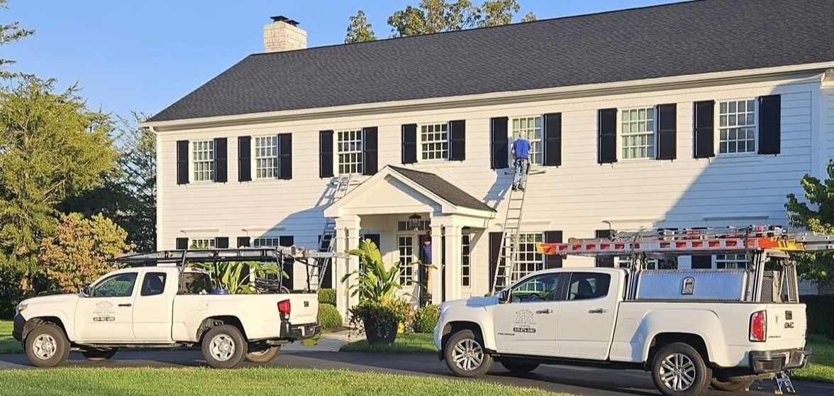 White house with black shutters, two white work trucks parked on a lawn, worker on a ladder.