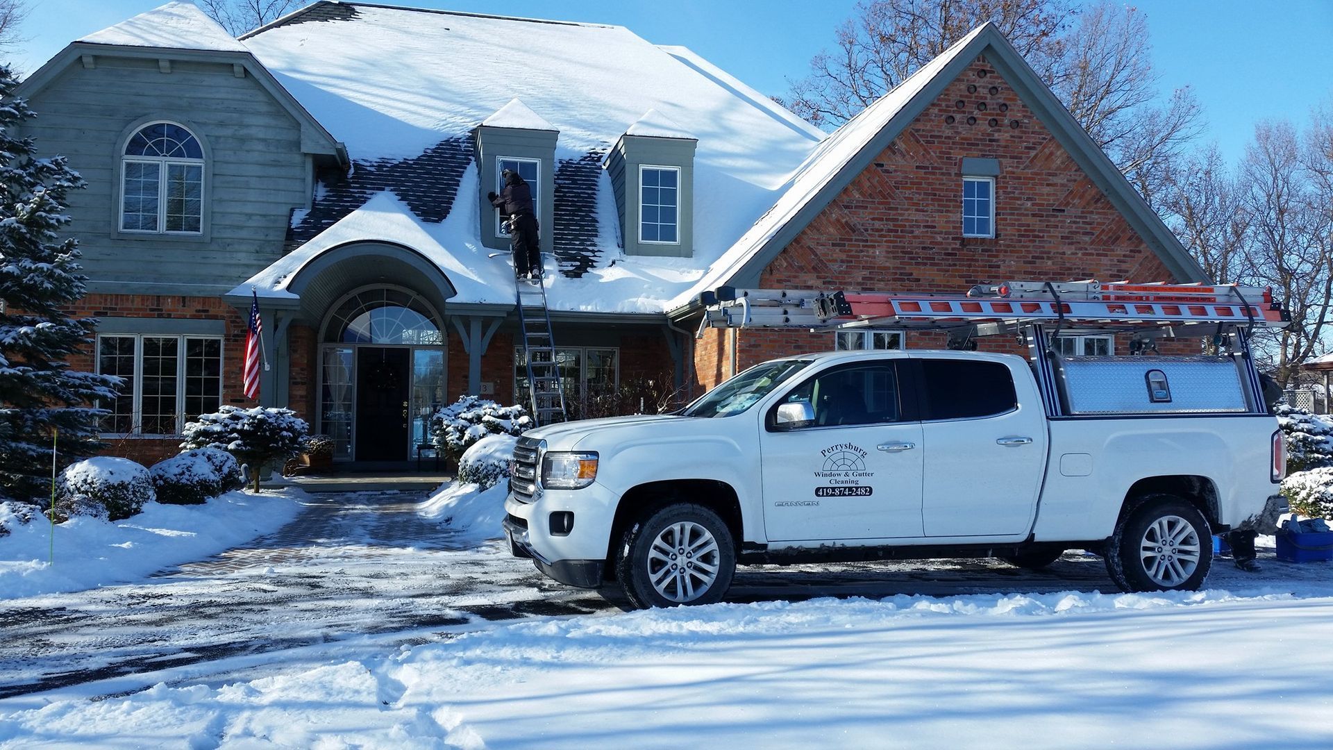White truck parked in front of a snow-covered house; worker on a ladder clearing snow from the roof.