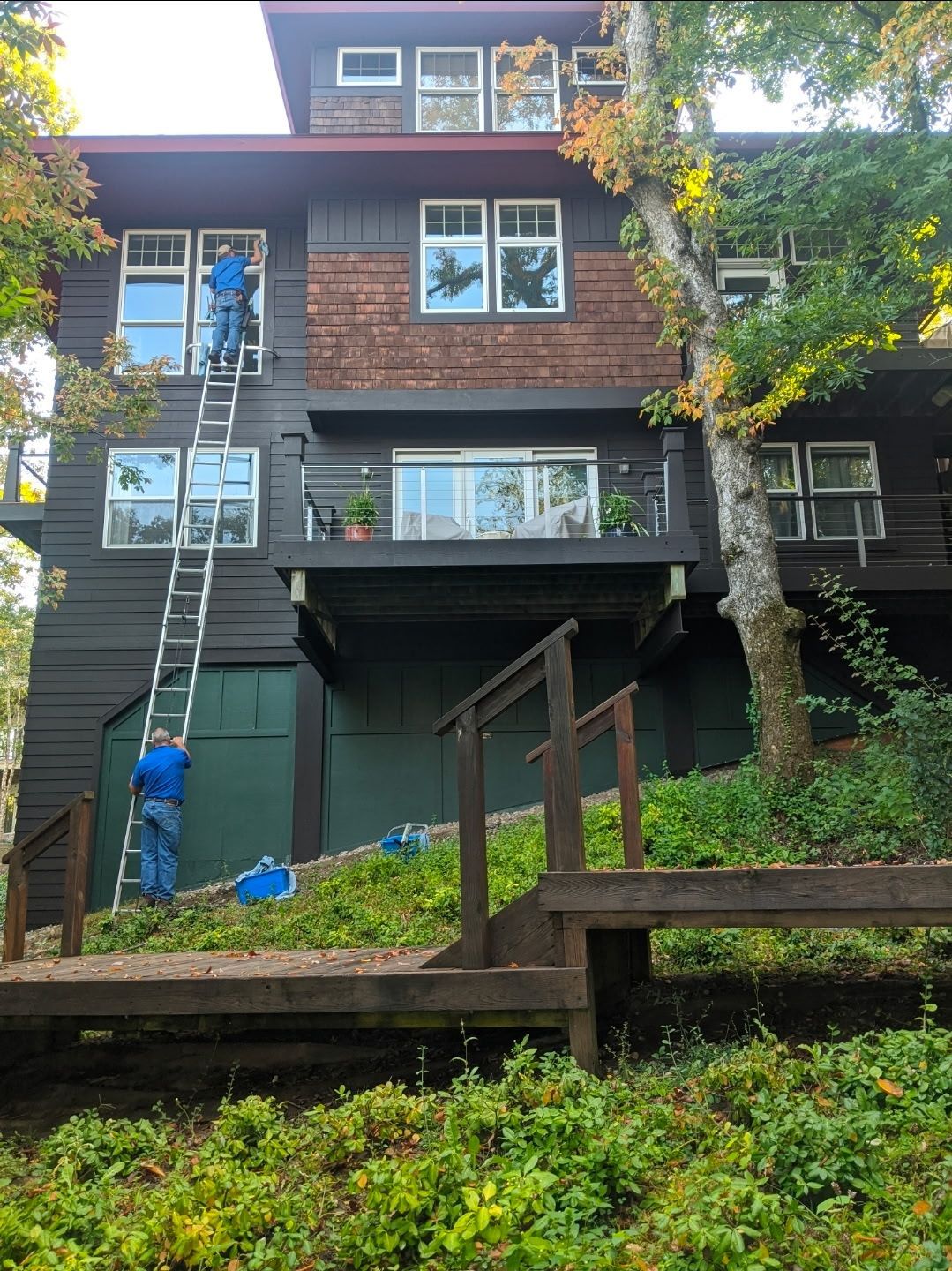 Two people painting a dark brown house, one on a ladder. House has a wooden deck and red shingle accents.