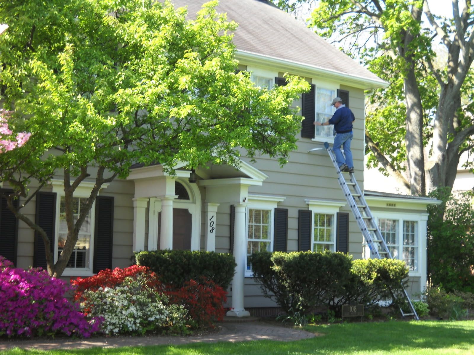 Person on ladder painting window of a two-story beige house. Lush green trees and colorful bushes in the yard.