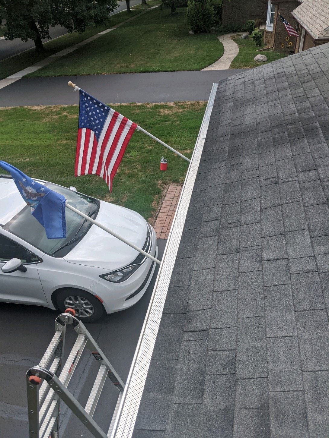 American flag and car with blue flag in a driveway next to a roof. Ladder is in view.