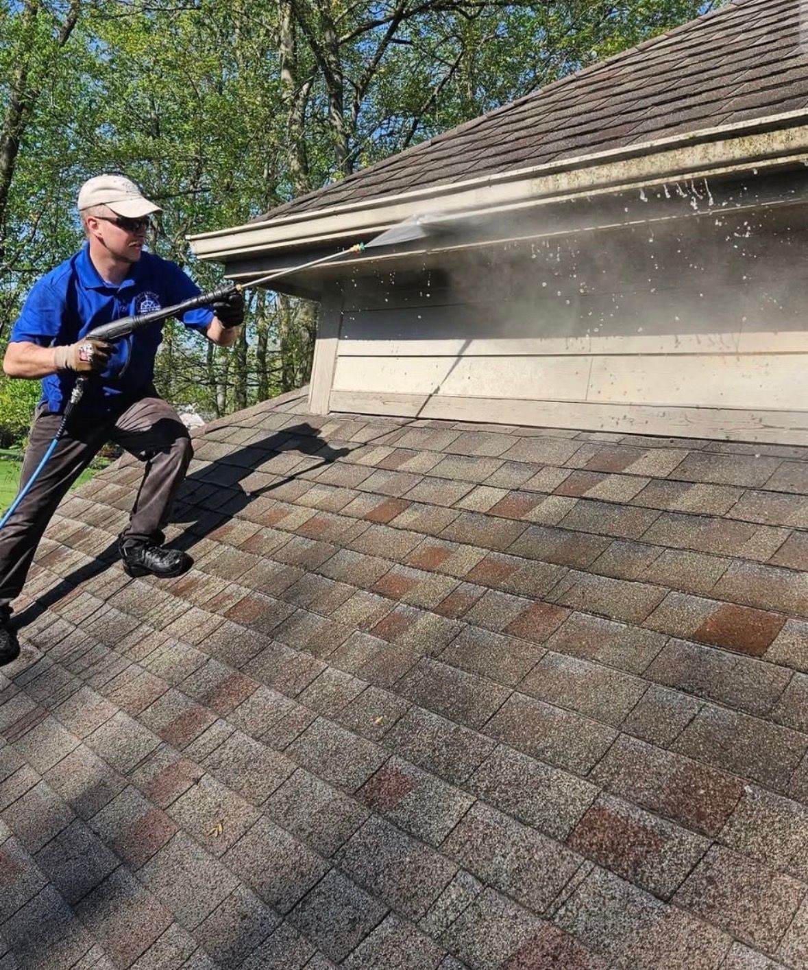 Worker pressure-washing a shingled roof near the gutter line with water spray visible