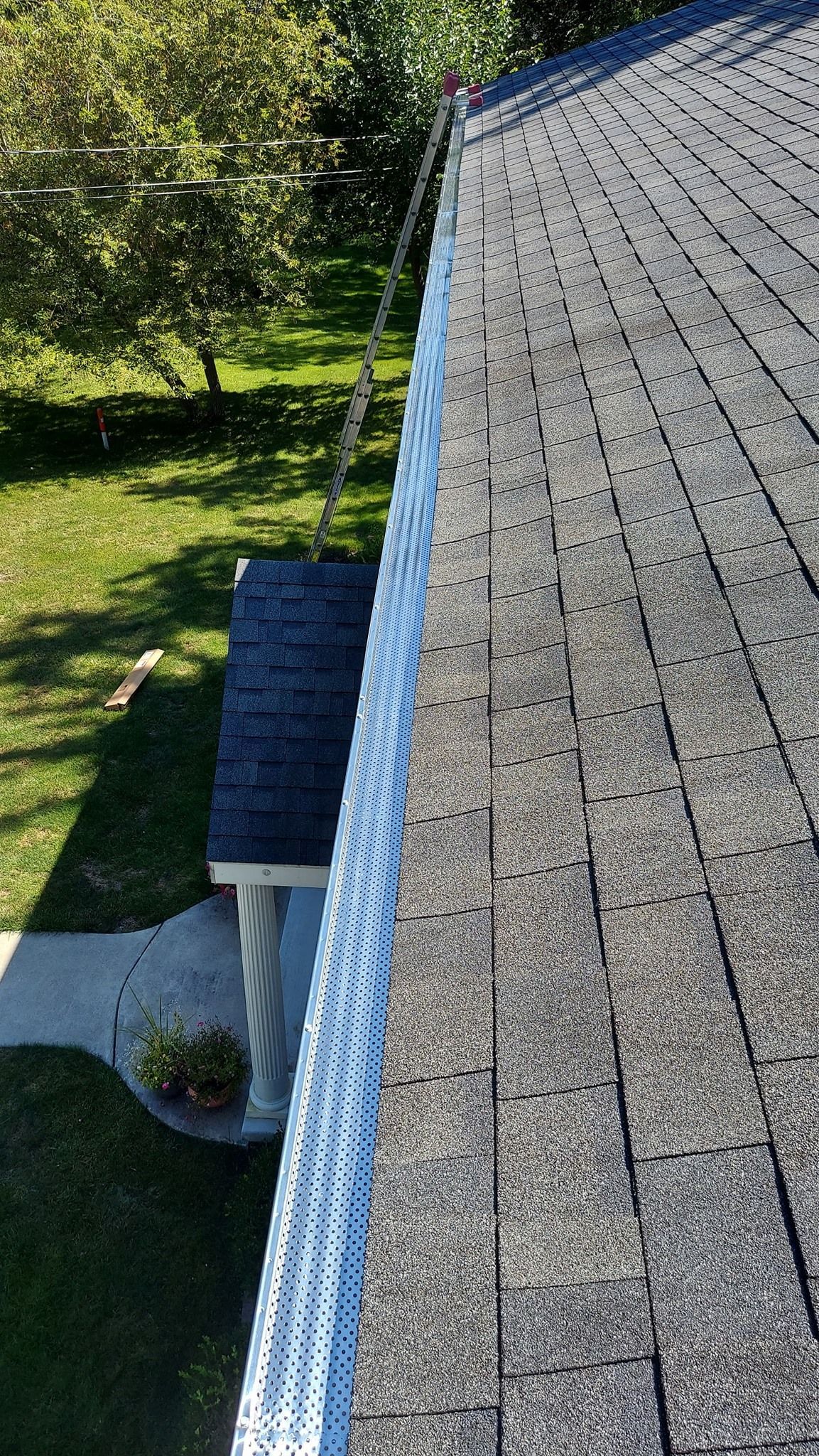 Overhead view of a roof with gray shingles and a metal gutter, with a green lawn and tree in the background.