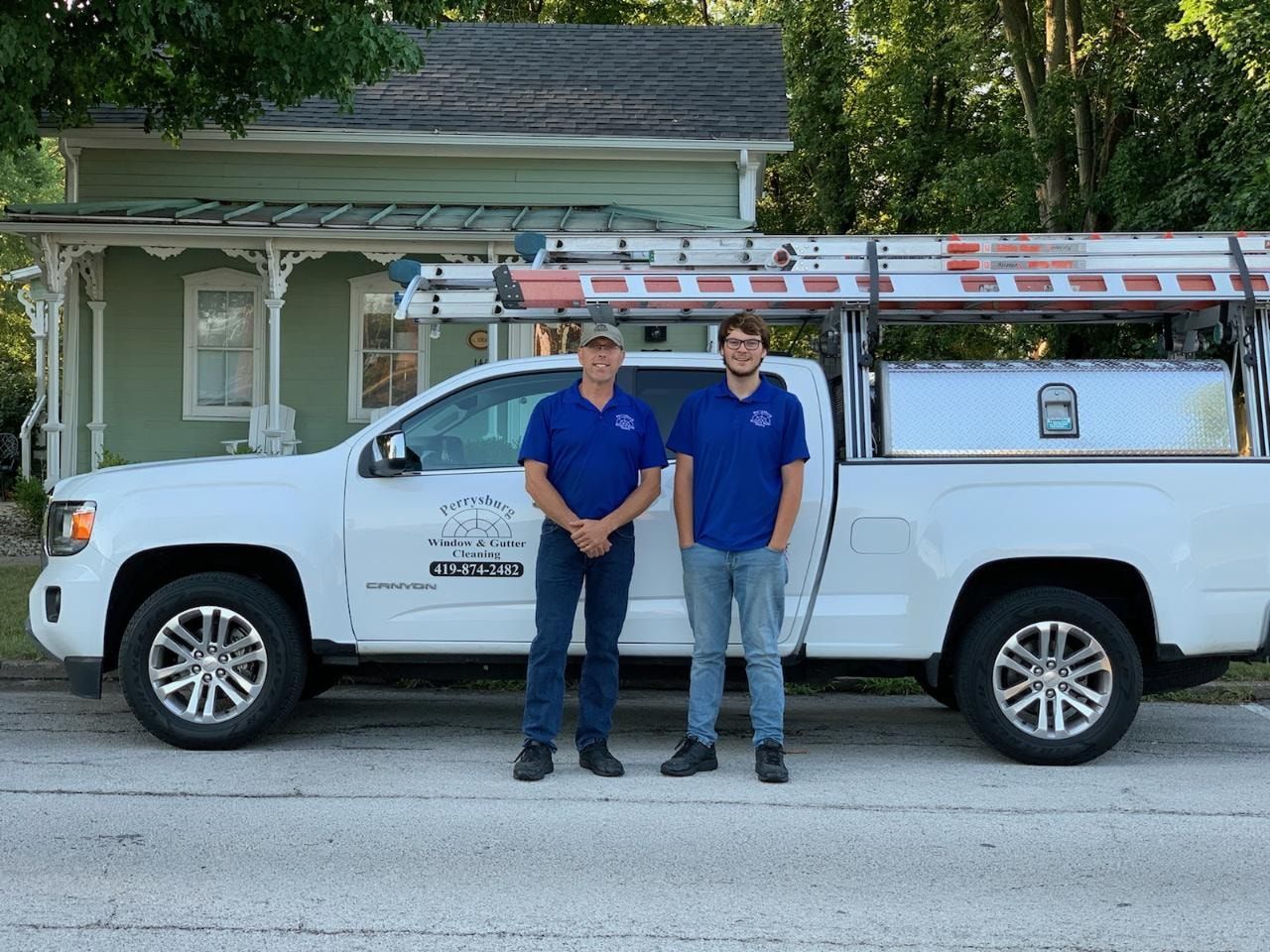 Two men in blue shirts stand beside a white truck with tools in front of a green house.