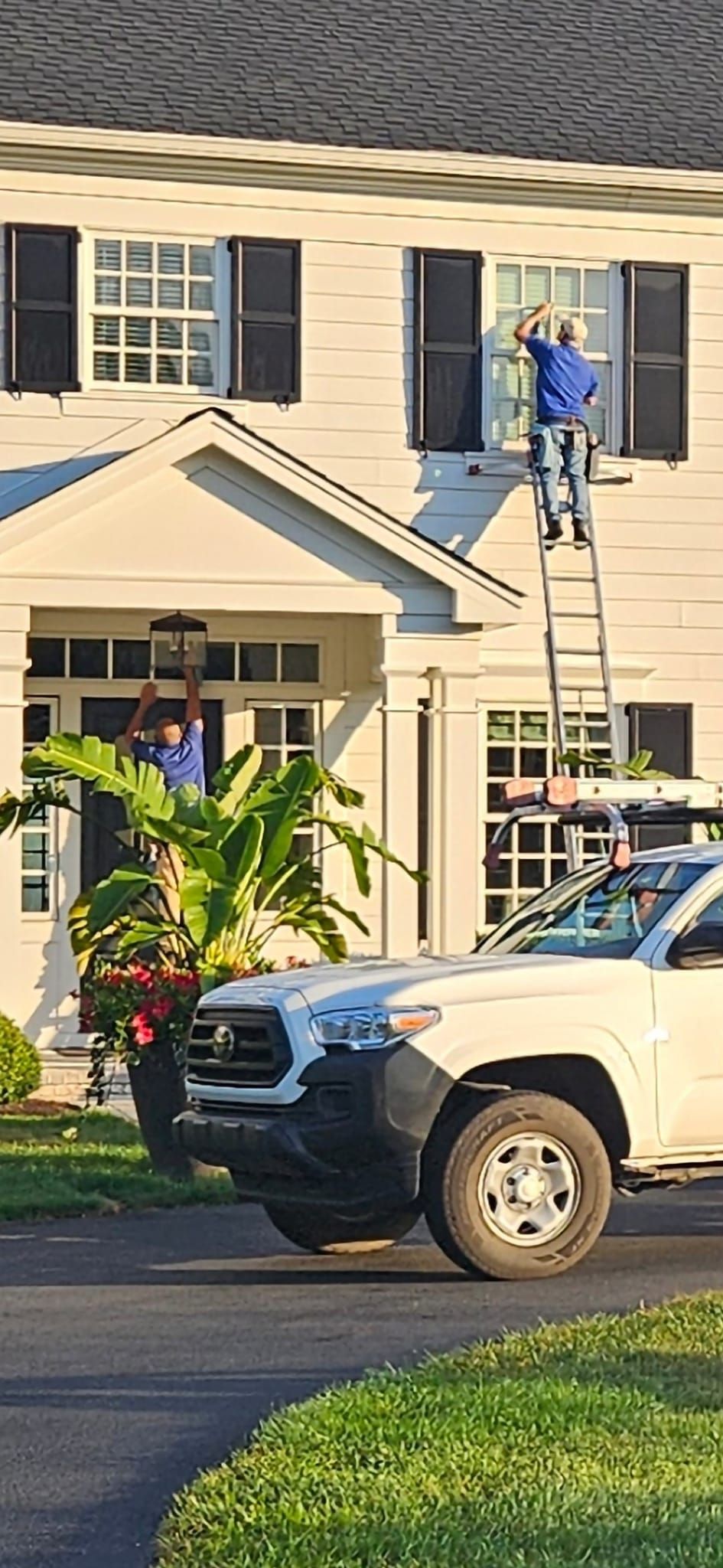 Two people cleaning windows on a white house, one on a ladder, and a truck parked out front.