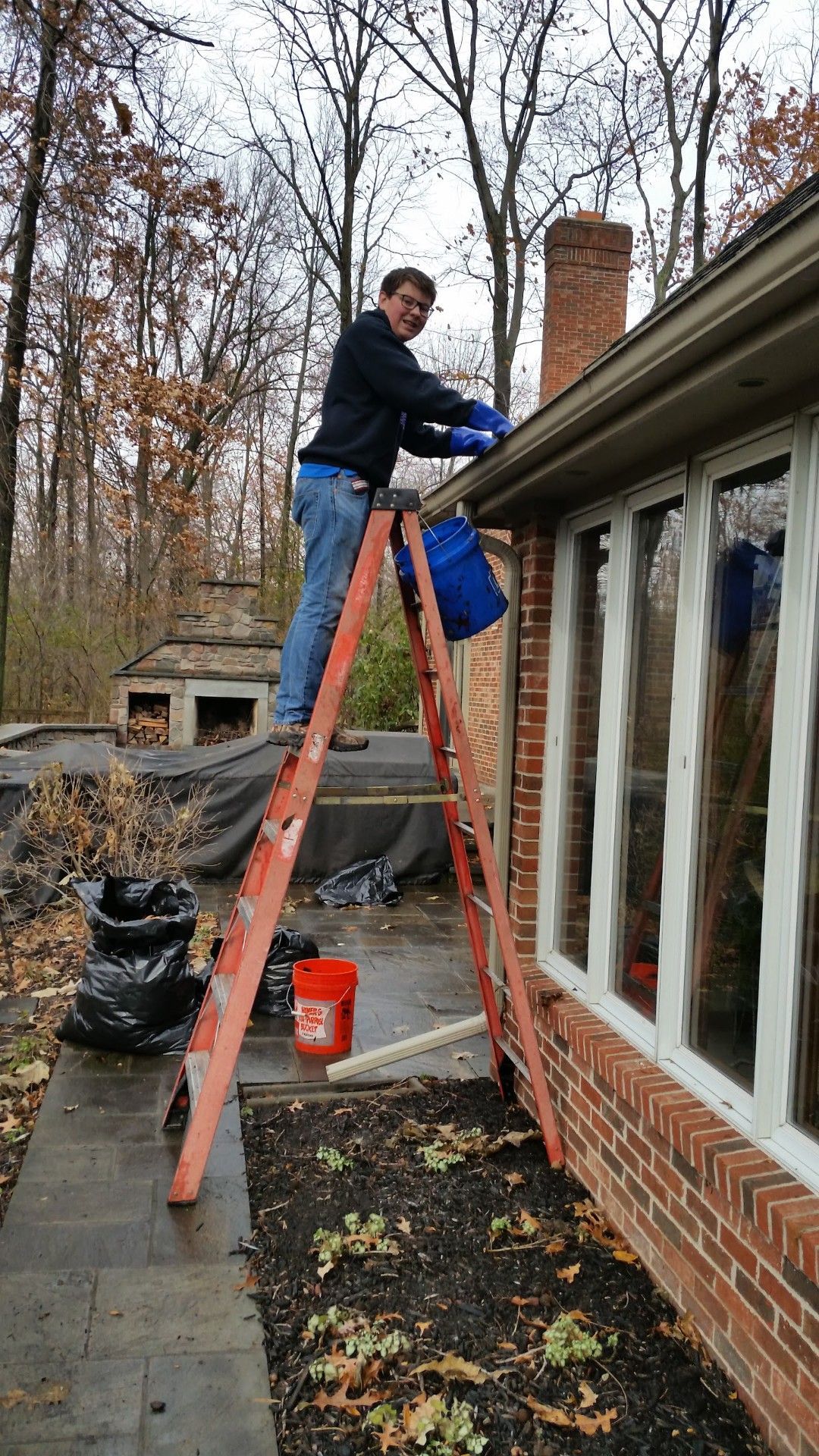 Man on a ladder cleaning a gutter near a brick building with trees in the background.