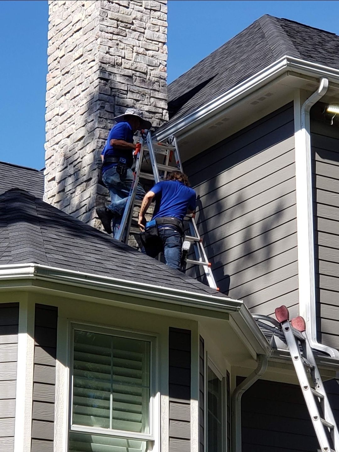 Two workers on ladders cleaning a chimney on a house with gray siding and dark roof.
