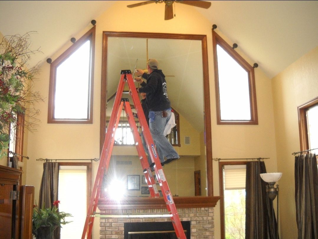 Person on a ladder installing a large mirror above a fireplace, flanked by windows.
